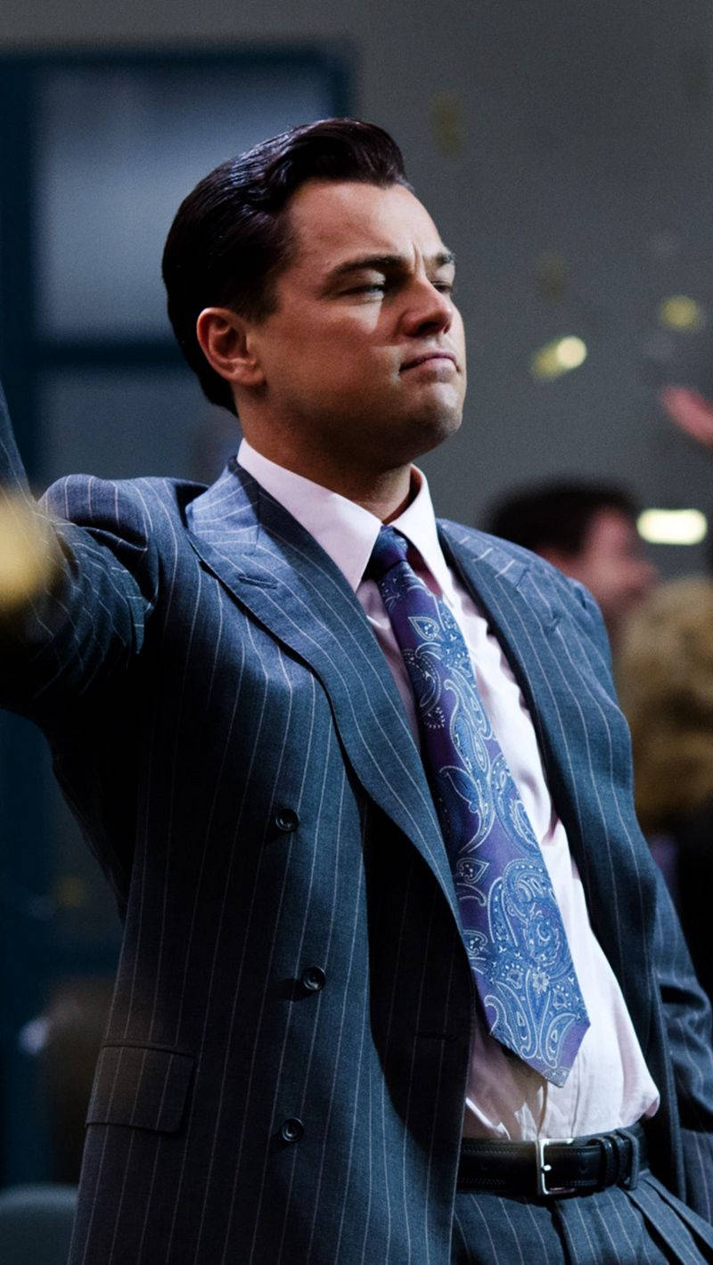 Dicaprio 桌布 壁紙 — 一名身穿細條紋西裝和佩斯利領帶的男子，在光線昏暗的房間裡舉起手臂，背景人物模糊。