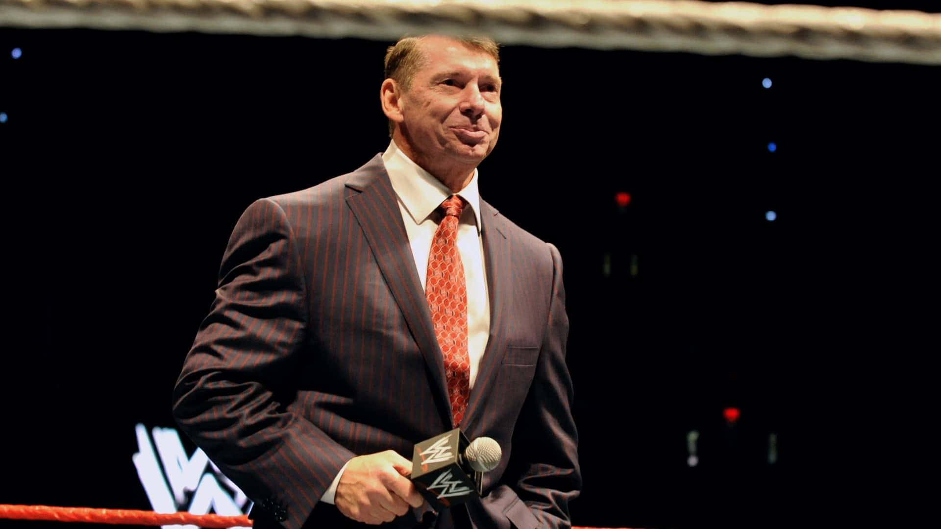 WWE 桌布 壁紙 — 一名身穿細條紋西裝和紅色領帶的男子在明亮的舞台燈光下自信地站在摔角擂台上，手持麥克風。