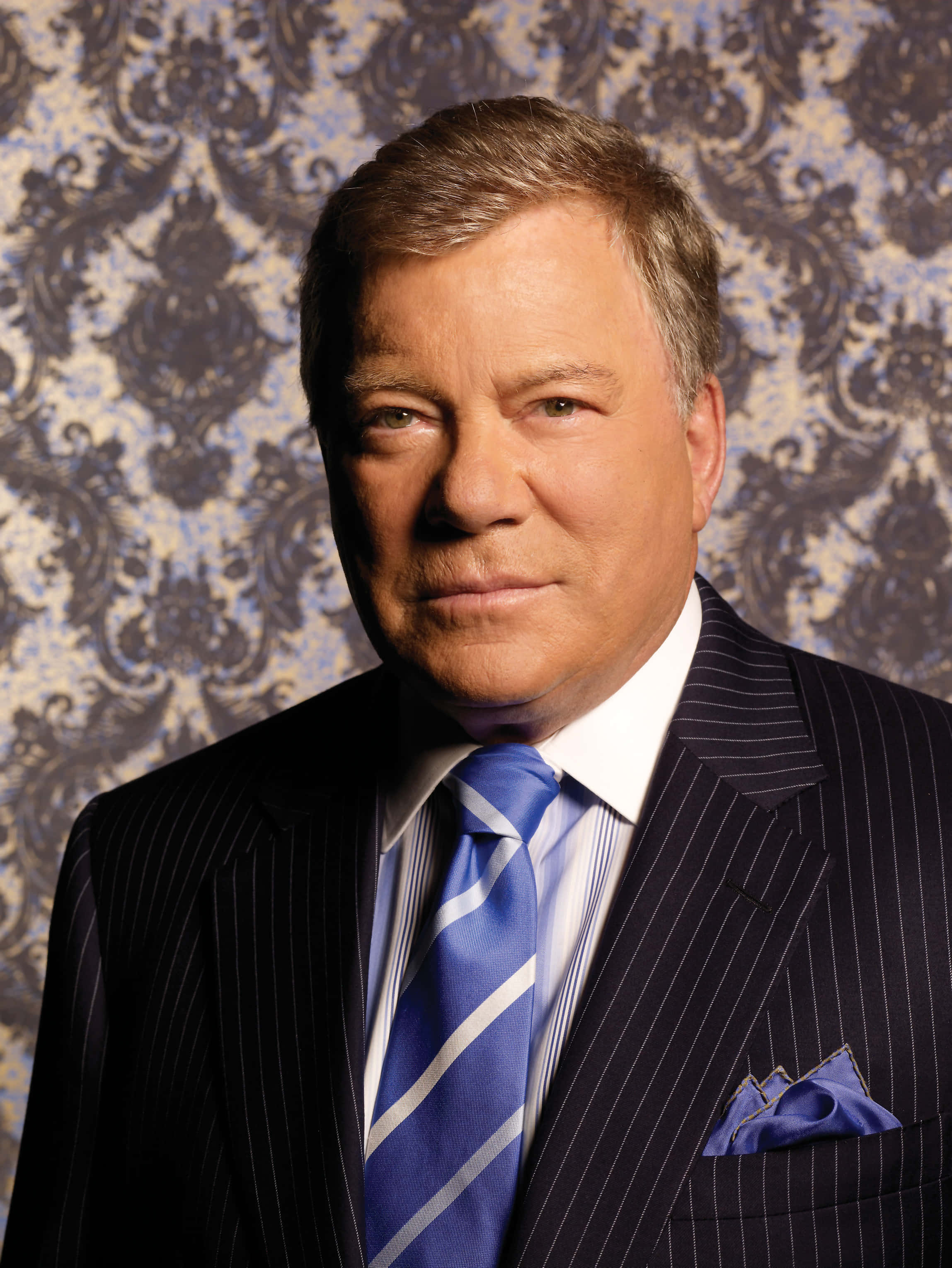 william shatner 桌布 壁紙 — 一名身穿細條紋西裝和藍色領帶的男子，表情嚴肅地站在華麗的錦緞壁紙前。