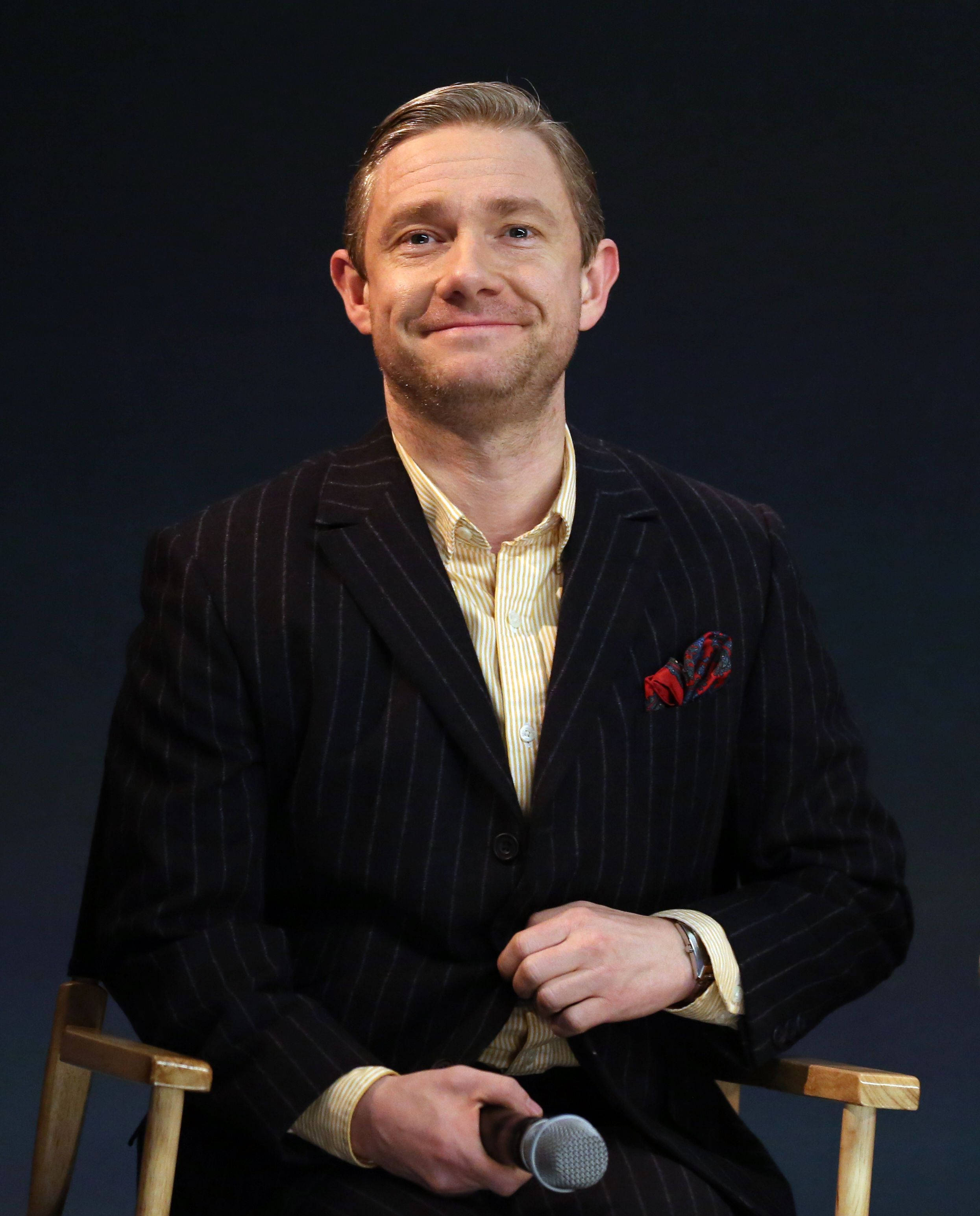 Martin Freeman 桌布 壁紙 — 一名身穿細條紋西裝的男子在深色背景下的導演椅上微笑著手持麥克風。