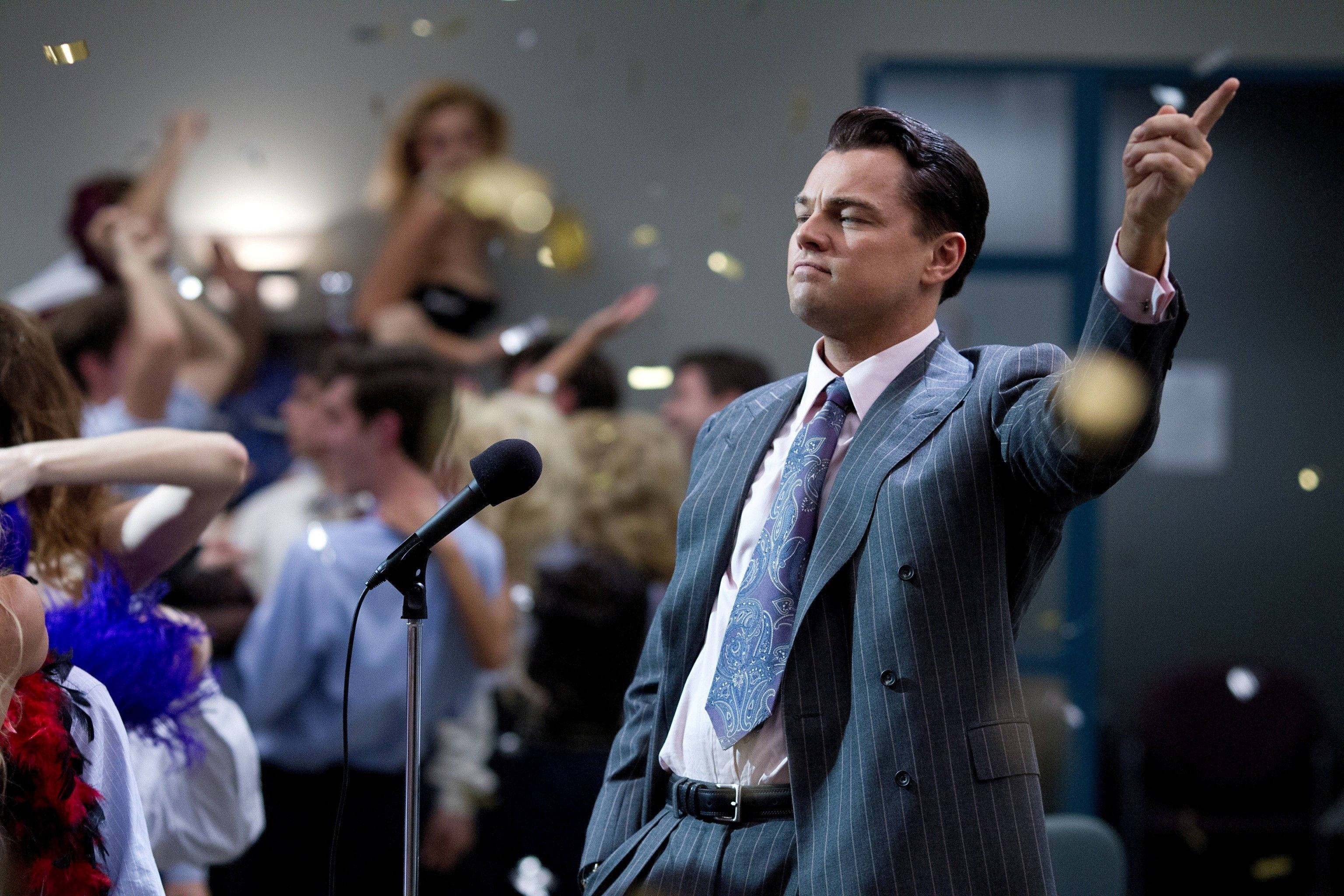 Wolf Of Wall Street 桌布 壁紙 — 一名身穿細條紋西裝的男子在熱鬧的派對中，於飄落的金色紙屑和模糊的賓客之間向上指。