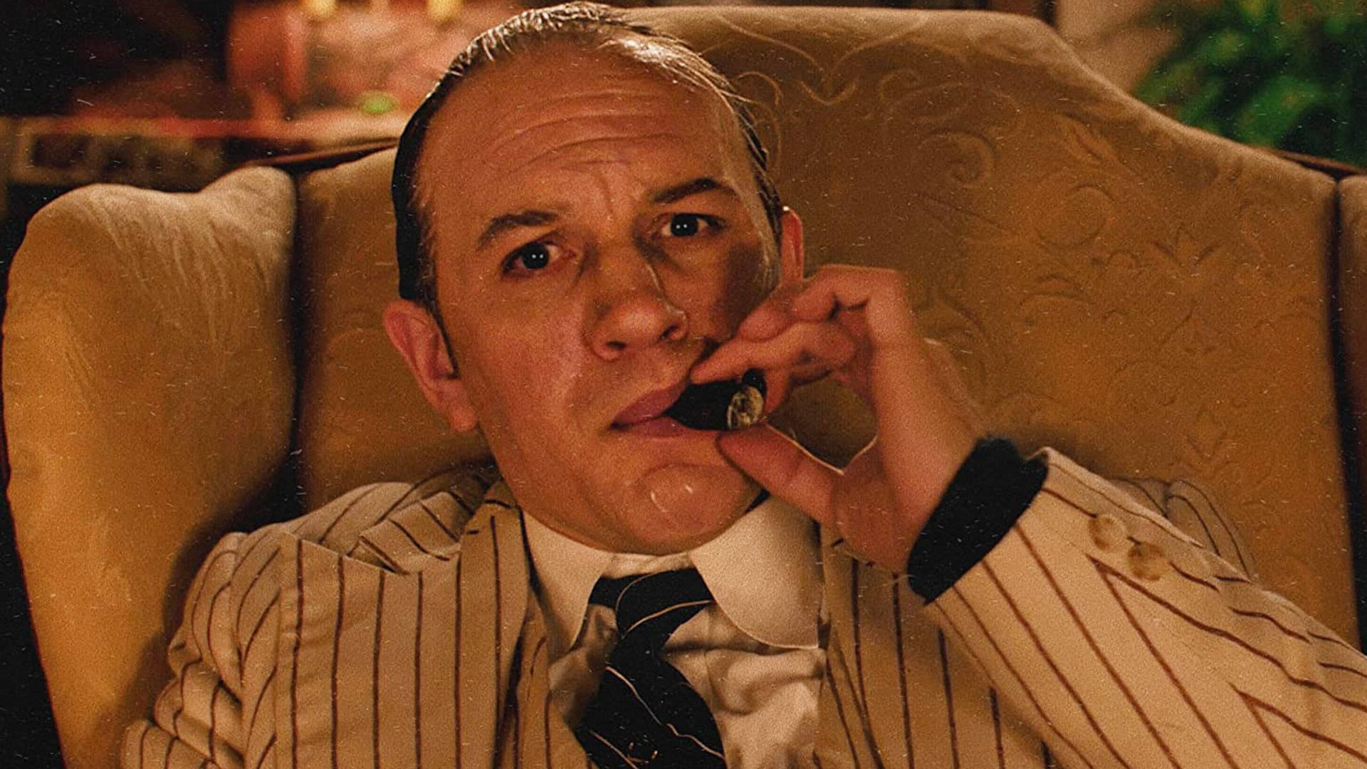 Capone 桌布 壁紙 — 一名身穿細條紋西裝的男子坐在豪華扶手椅上，在溫暖的金色燈光下將雪茄放在唇邊。