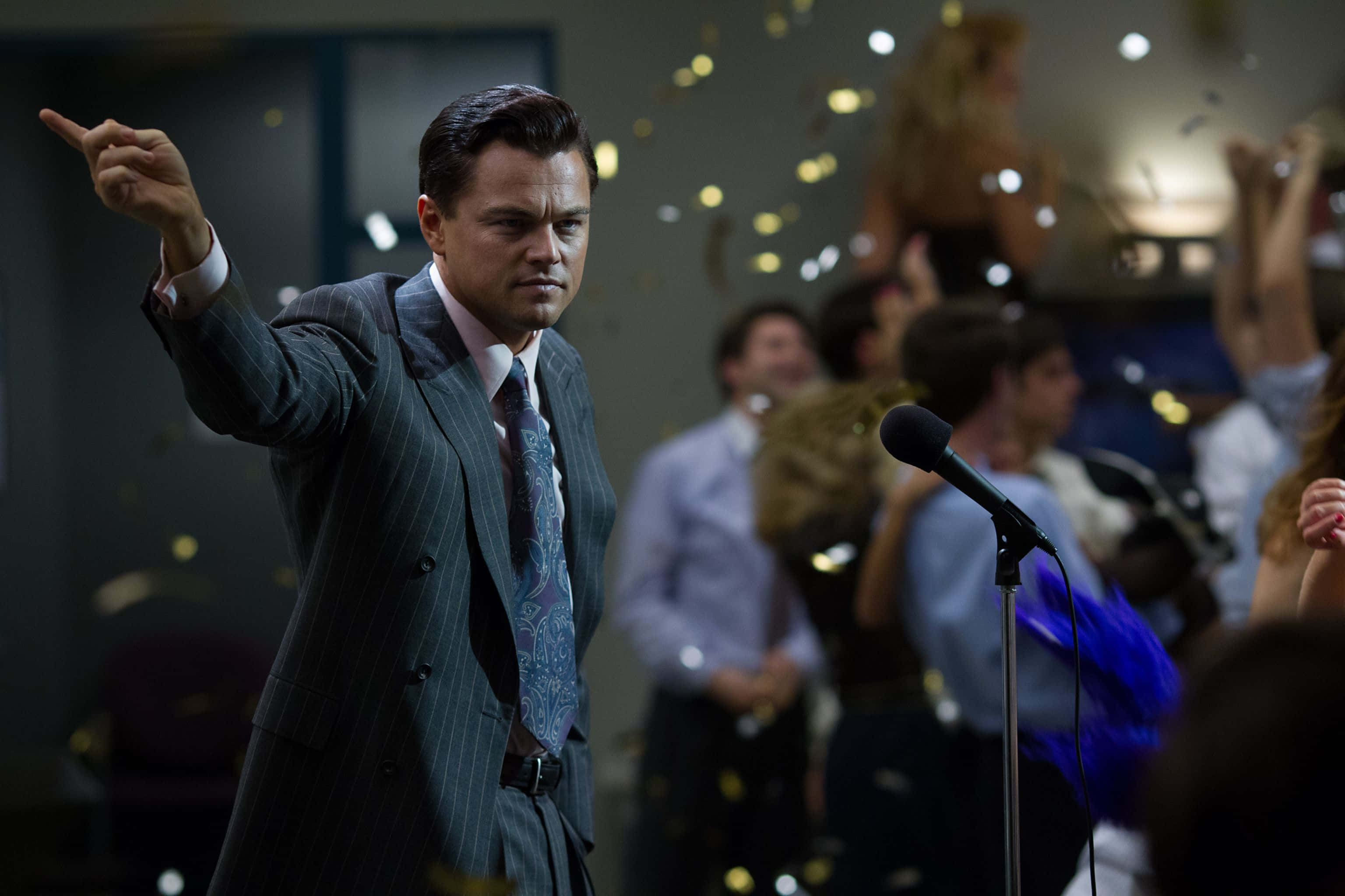Wolf Of Wall Street 桌布 壁紙 — 一名身穿細條紋西裝的男子權威地指向人群，金色紙屑在他周圍飄落。