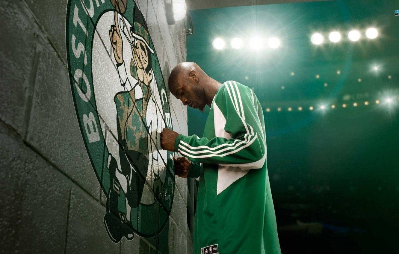 Kevin Garnett 桌布 壁紙 — 一名身穿綠白球衣的籃球運動員在明亮的場館燈光下，在混凝土牆上的波士頓塞爾提克標誌前恭敬地鞠躬。
