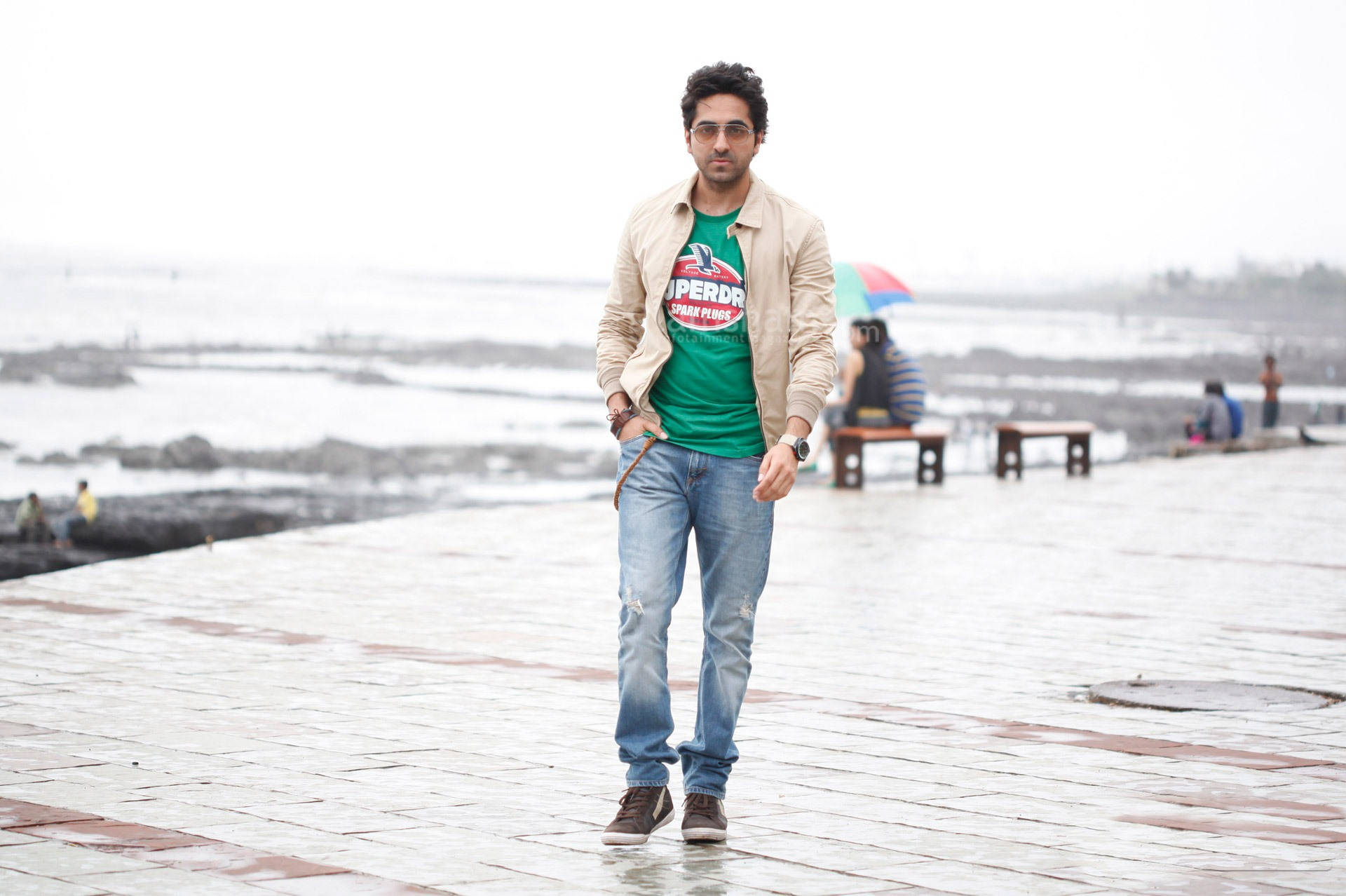 ayushmann khurrana 桌布 壁紙 — 一名身穿綠色T恤和米色外套的男子在陰天自信地走在濕漉漉的海濱長廊上。