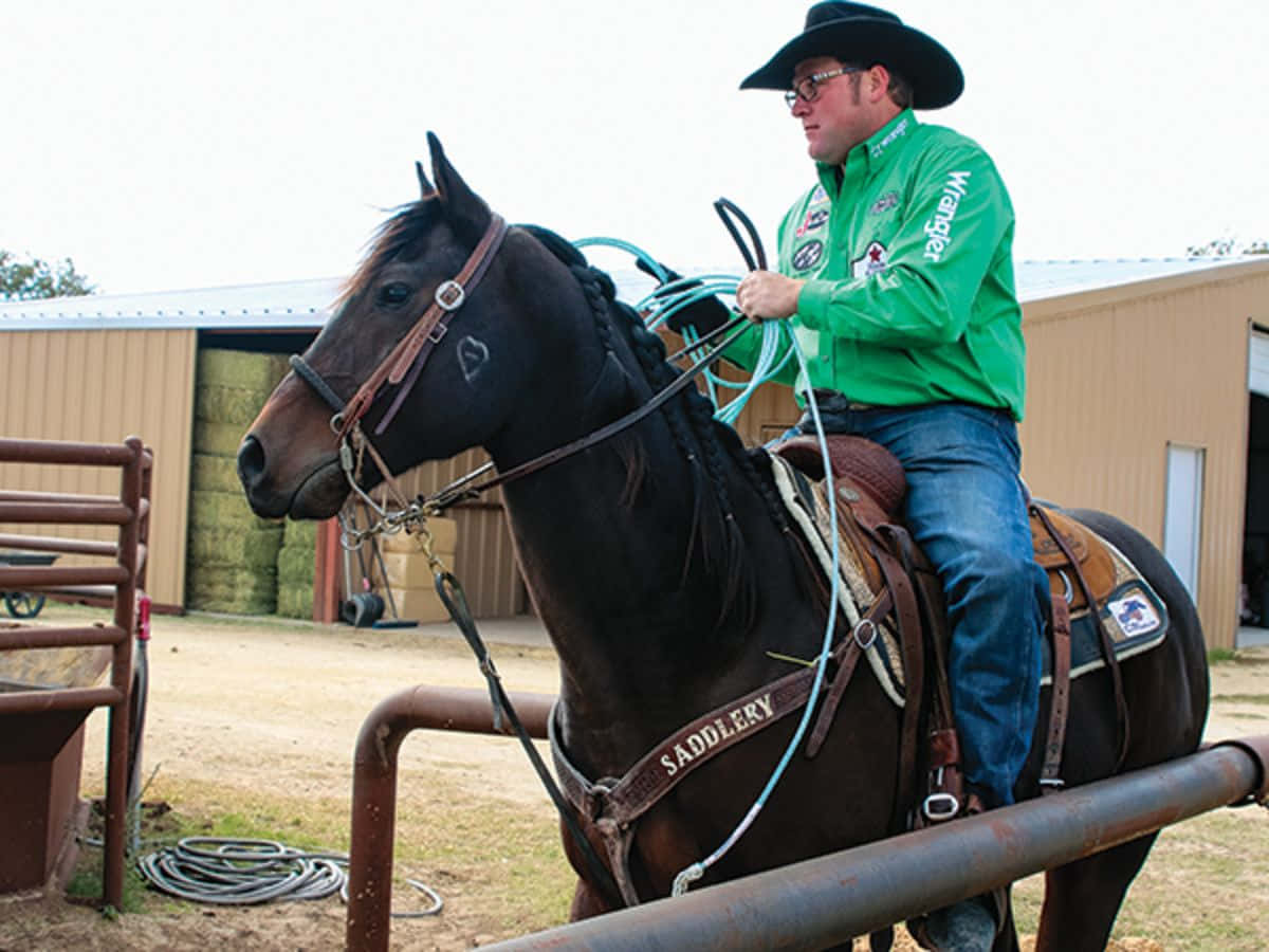 Roping 桌布 壁紙 — 一名身穿綠色Wrangler襯衫、戴著牛仔帽的男子，坐在深棕色馬上，手拿捲起的繩索，靠近一座質樸的穀倉。