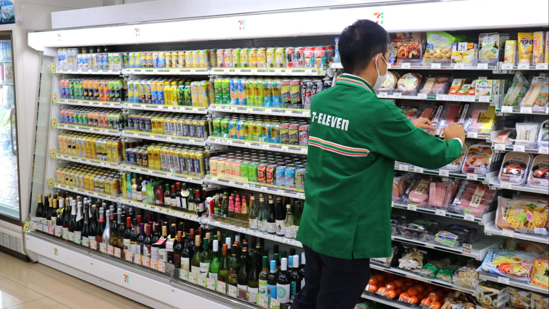 7-ELEVEN 桌布 壁紙 — 一名身穿綠色制服的7-eleven員工在光線明亮的便利商店貨架上補充冷藏飲品和即食餐點。