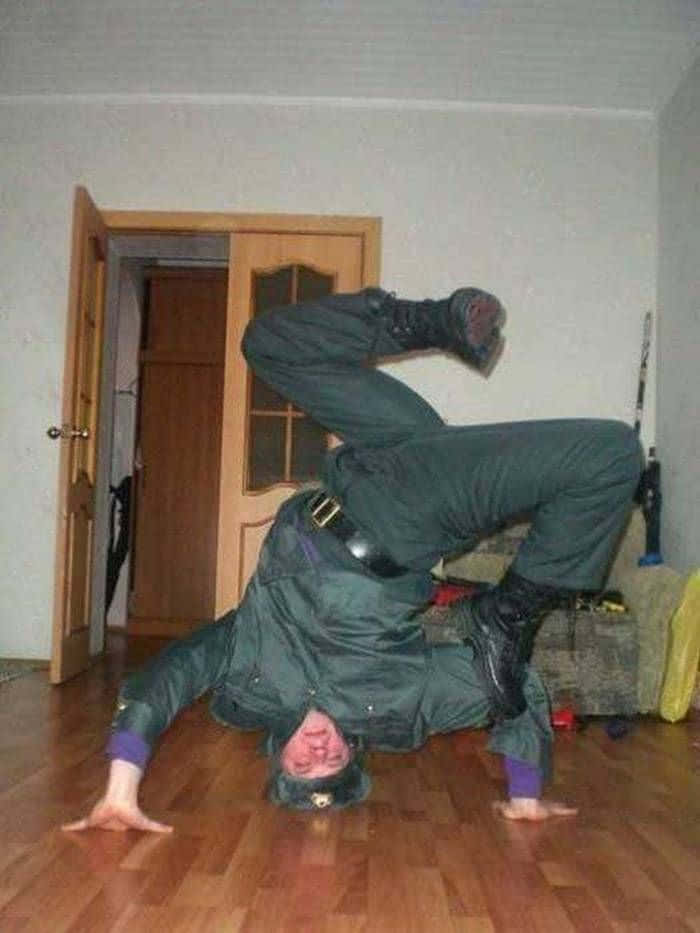 搞笑 桌布 壁紙 — 一名身穿綠色制服的人士在一個雜亂的室內房間中，在木地板上做分腿倒立。