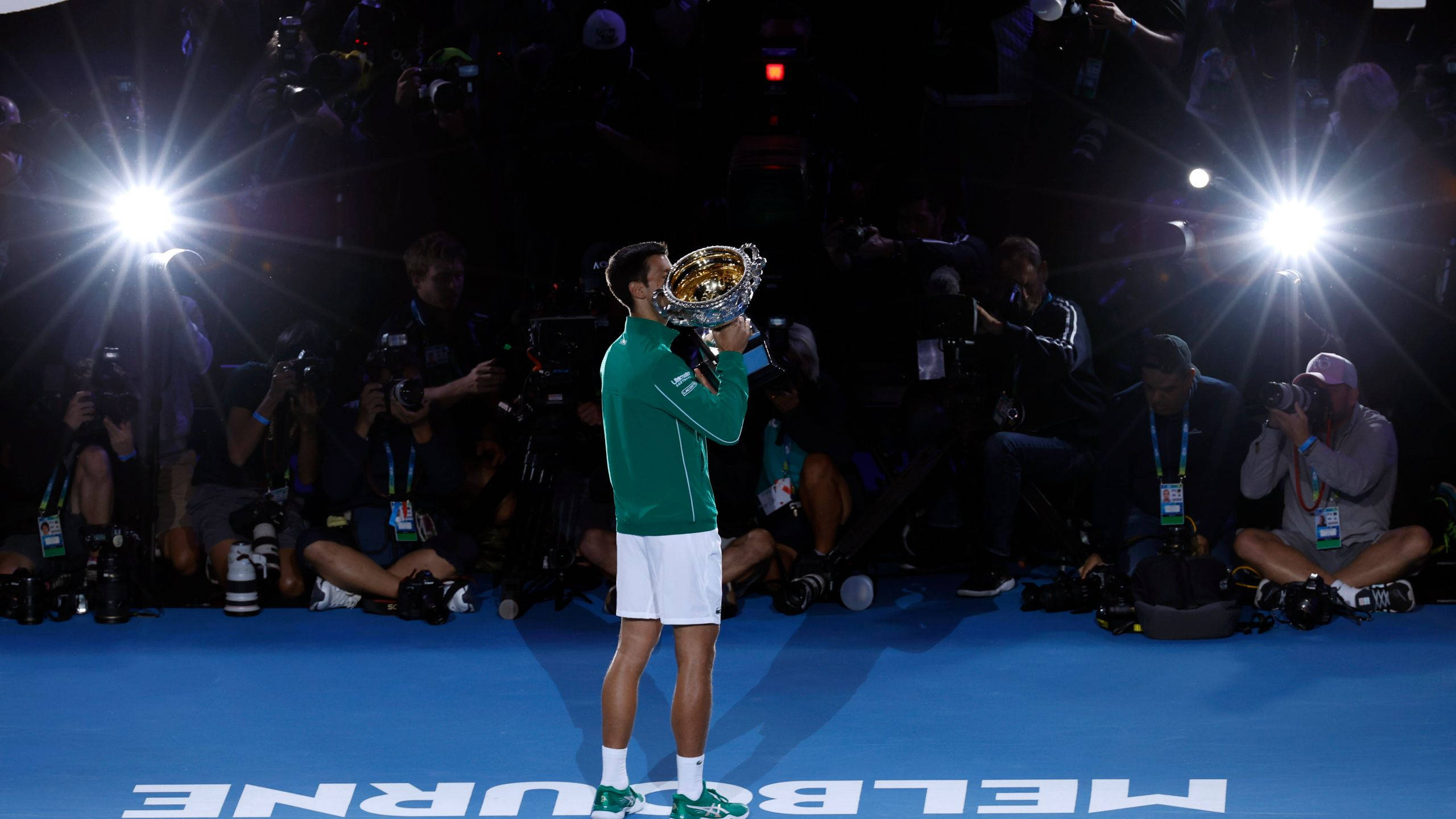 Australian Open 桌布 壁紙 — 一名身穿綠色和白色球衣的網球選手站在藍色球場上高舉獎盃，周圍的攝影師閃光燈不斷。