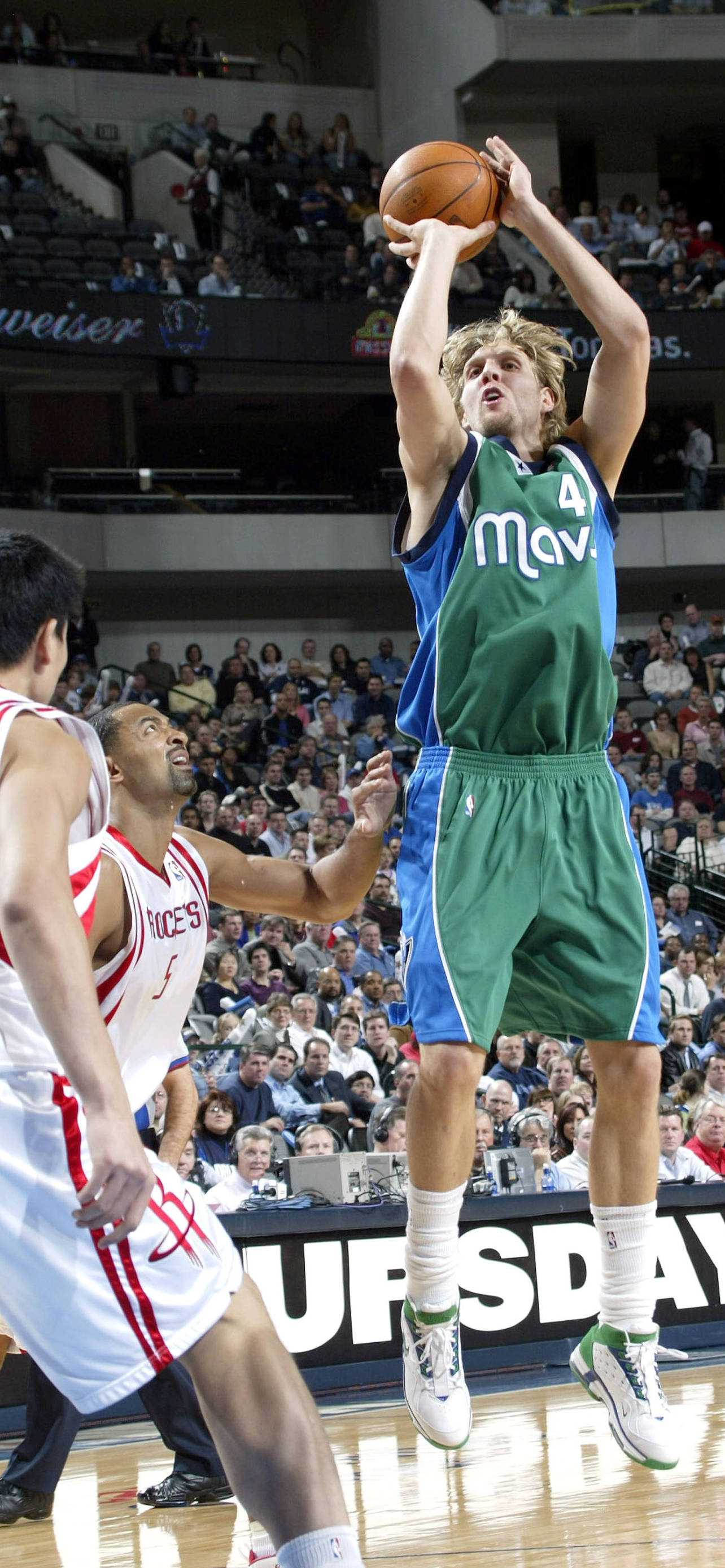 Dirk Nowitzki 桌布 壁紙 — 一名身穿綠色和藍色球衣的籃球運動員高高躍起投籃，而一名身穿白色的防守球員試圖封阻。