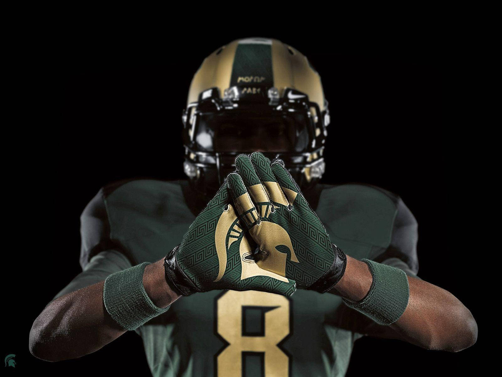 Michigan State Spartans 桌布 壁紙 — 一名身穿綠色和金色制服、戴著頭盔和手套的美式足球員在黑色背景前將雙手放在臉前。