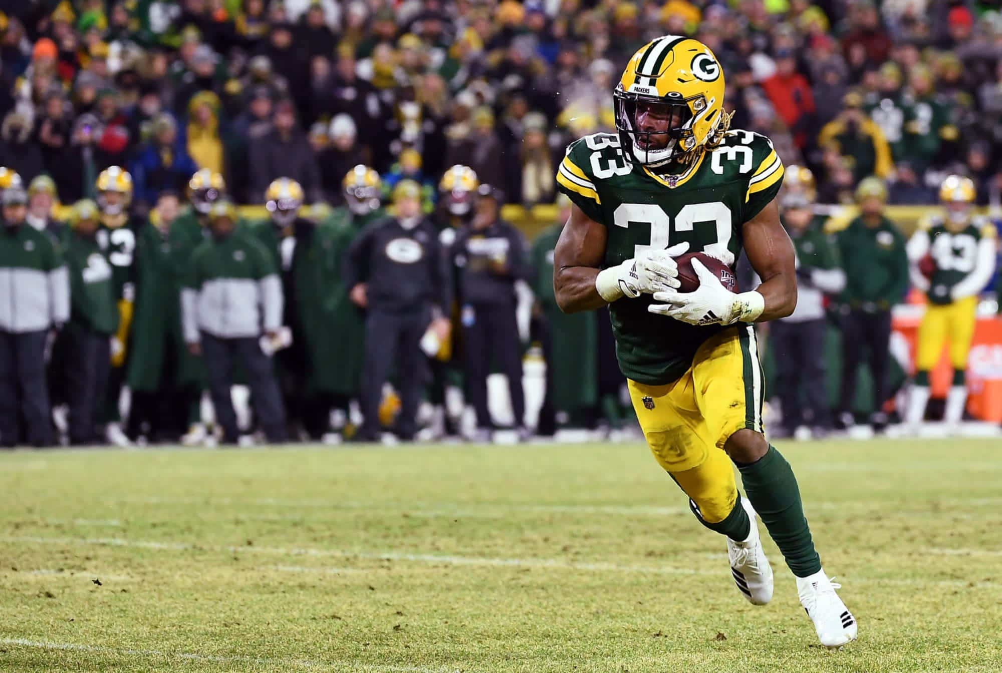 Aaron Jones 桌布 壁紙 — 一名身穿綠色和金色球衣的綠灣包裝工美式足球員在夜間比賽中，在球場燈光下帶球衝刺。