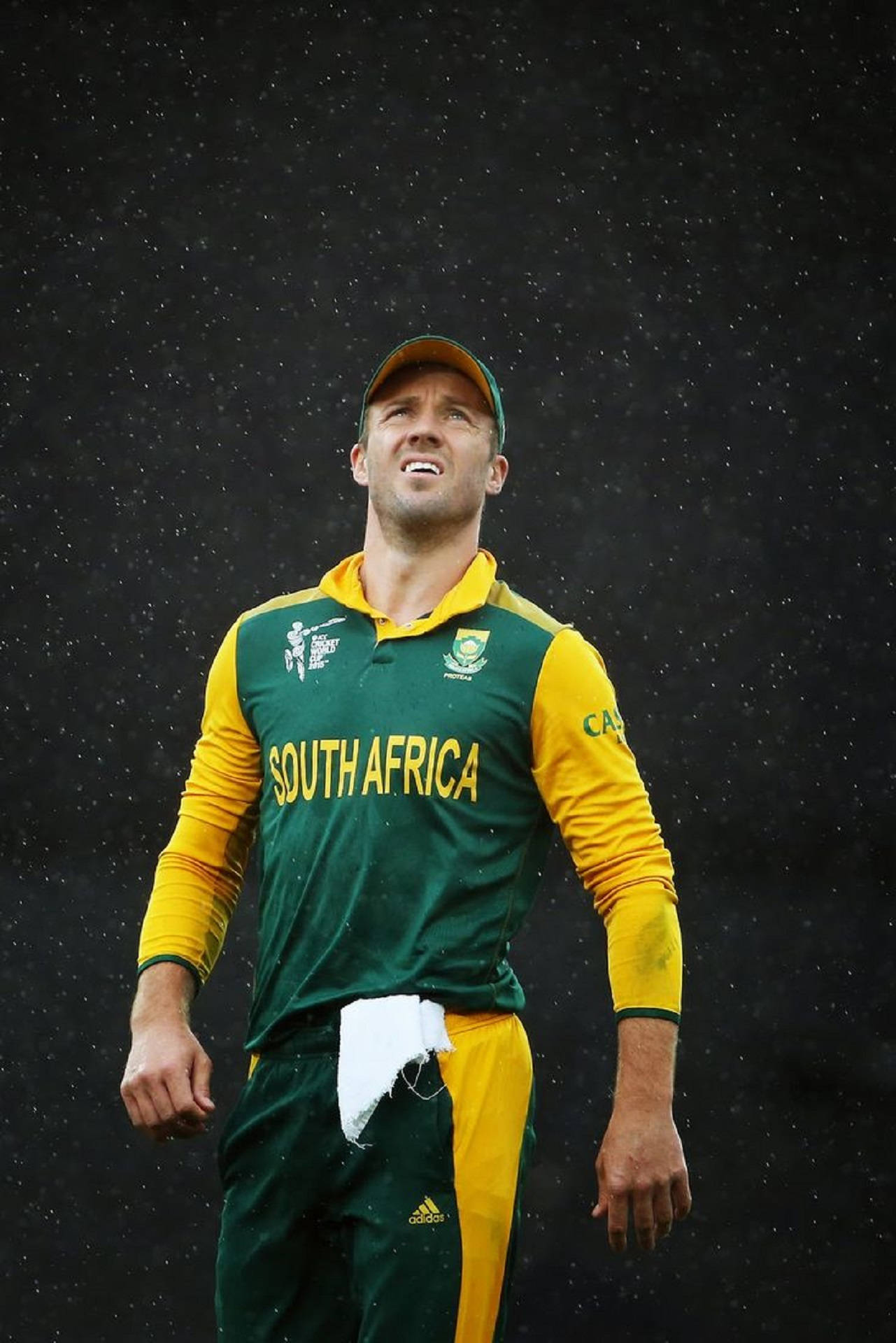 south africa cricket 桌布 壁紙 — 一名身穿綠色和黃色球衣的南非板球運動員在雨中站立，表情堅定，背景昏暗。