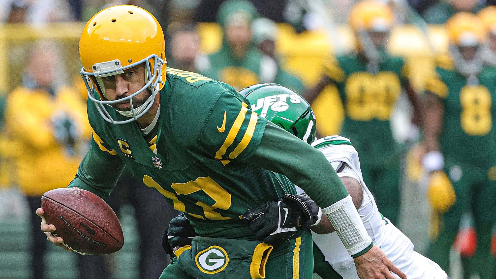 Aaron Rodgers 桌布 壁紙 — 一名身穿綠色和黃色球衣的綠灣包裝工四分衛帶著橄欖球奔跑，同時被紐約噴射機防守球員擒抱。