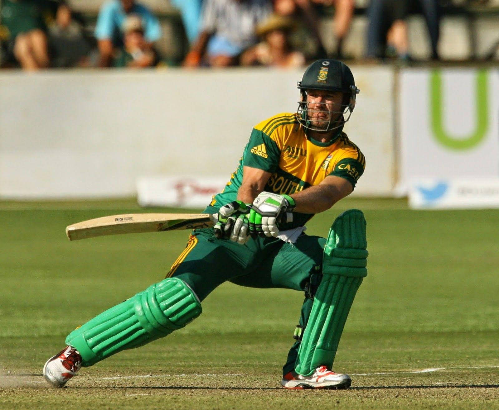 Ab De Villiers 桌布 壁紙 — 一名身穿綠色和黃色裝備的板球擊球手在陽光明媚的場地上低蹲準備擊球。