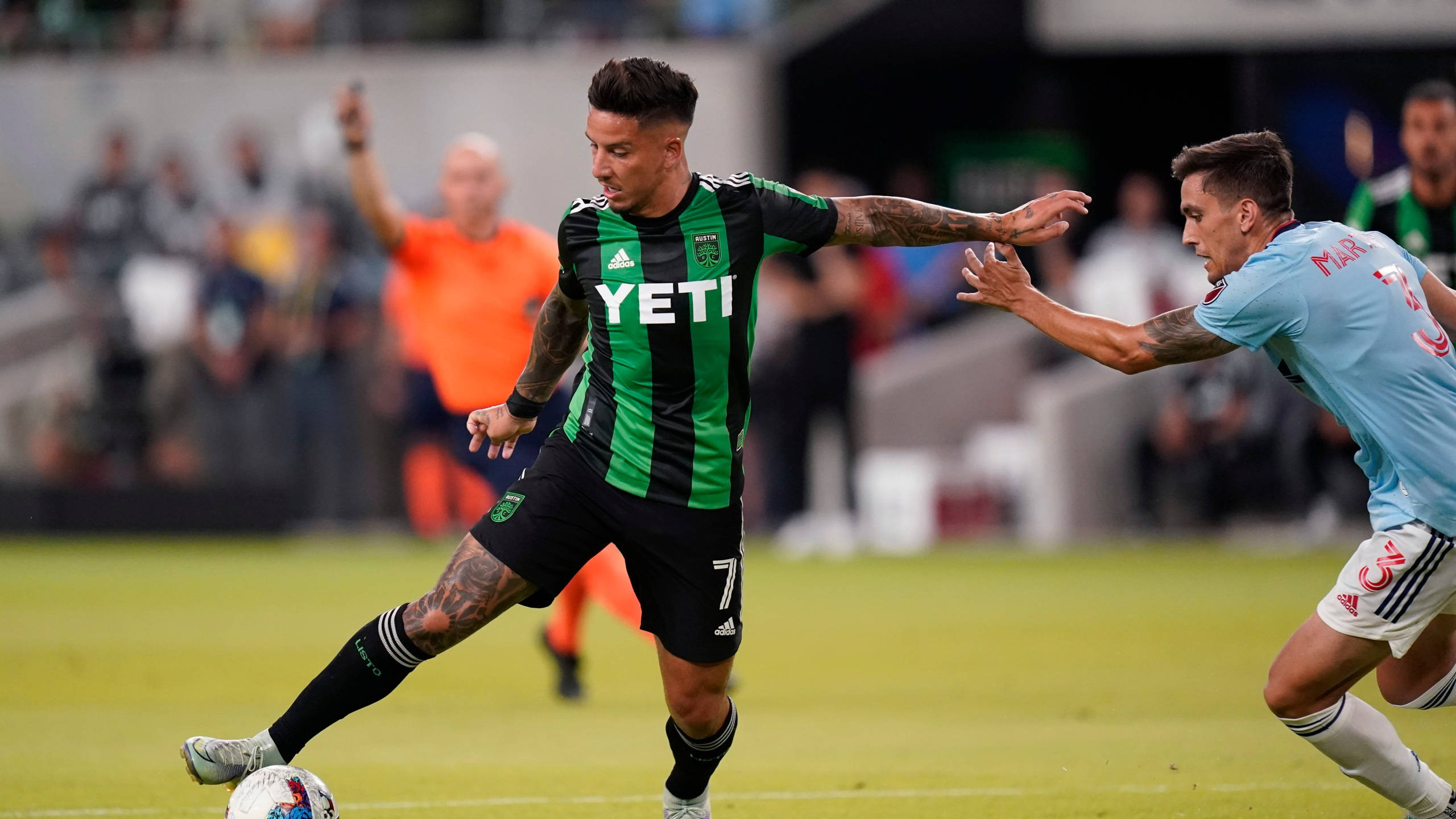 Austin Fc 桌布 壁紙 — 一名身穿綠色和黑色橫條球衣的足球員在草地上盤球，同時被一名身穿淺藍色球衣的對手攔截。