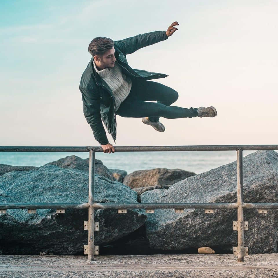jump 桌布 壁紙 — 一名身穿綠色外套的年輕男子動態地跳過金屬欄杆，身後是海洋和岩石。