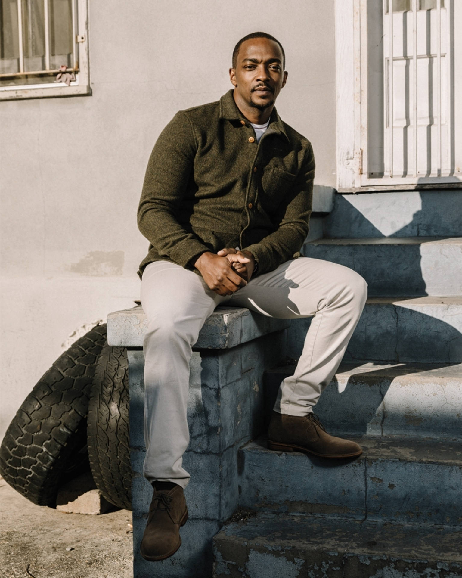 Anthony Mackie 桌布 壁紙 — 一名身穿綠色外套的男子在明亮的陽光下，平靜地坐在一個大輪胎旁邊的磨損藍色台階上。