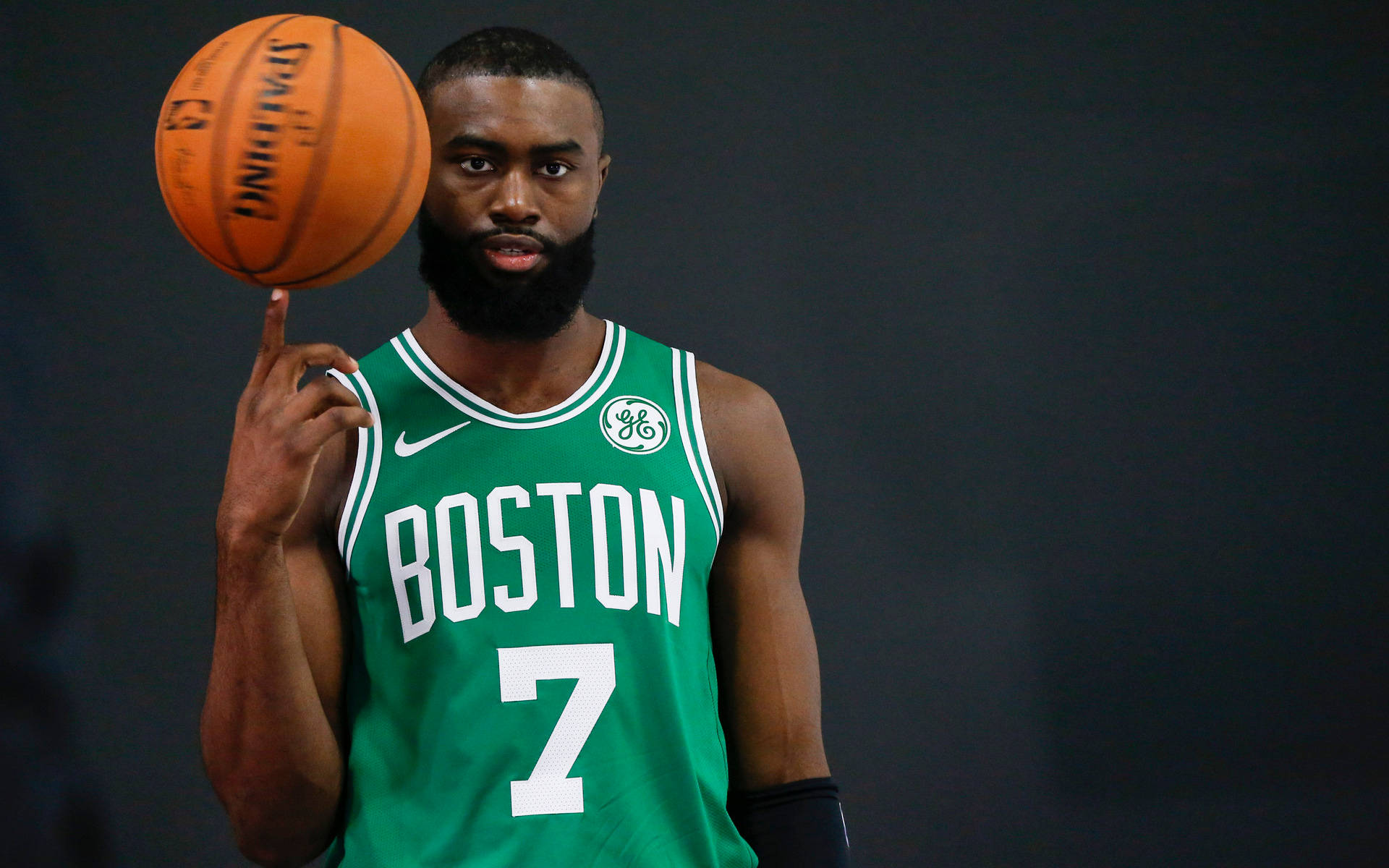 Jaylen Brown 桌布 壁紙 — 一名身穿綠色波士頓球衣的籃球運動員在深色背景下將橙色 Spalding 球平衡在指尖上。