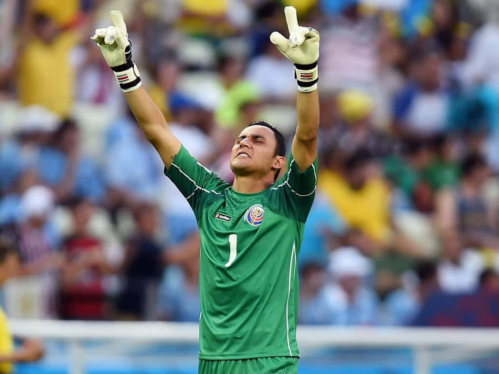 Keylor Navas 桌布 壁紙 — 一名身穿綠色球衣的足球守門員，在模糊的體育場人群背景下，高舉戴著手套的雙手慶祝勝利。