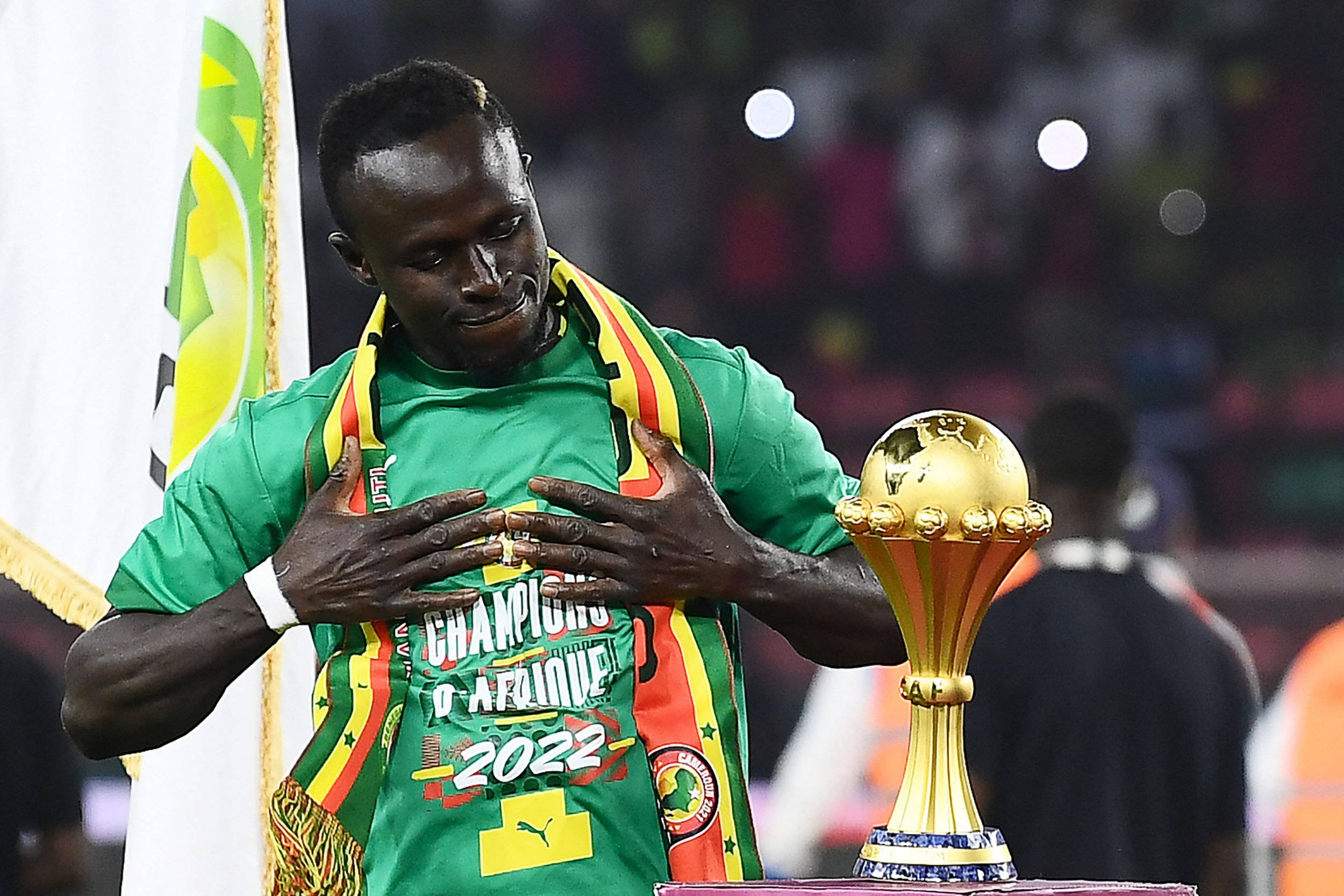 Senegal 桌布 壁紙 — 一名身穿綠色球衣的足球運動員在明亮燈光下的體育場內，雙手放在心口上捧著金色獎盃。