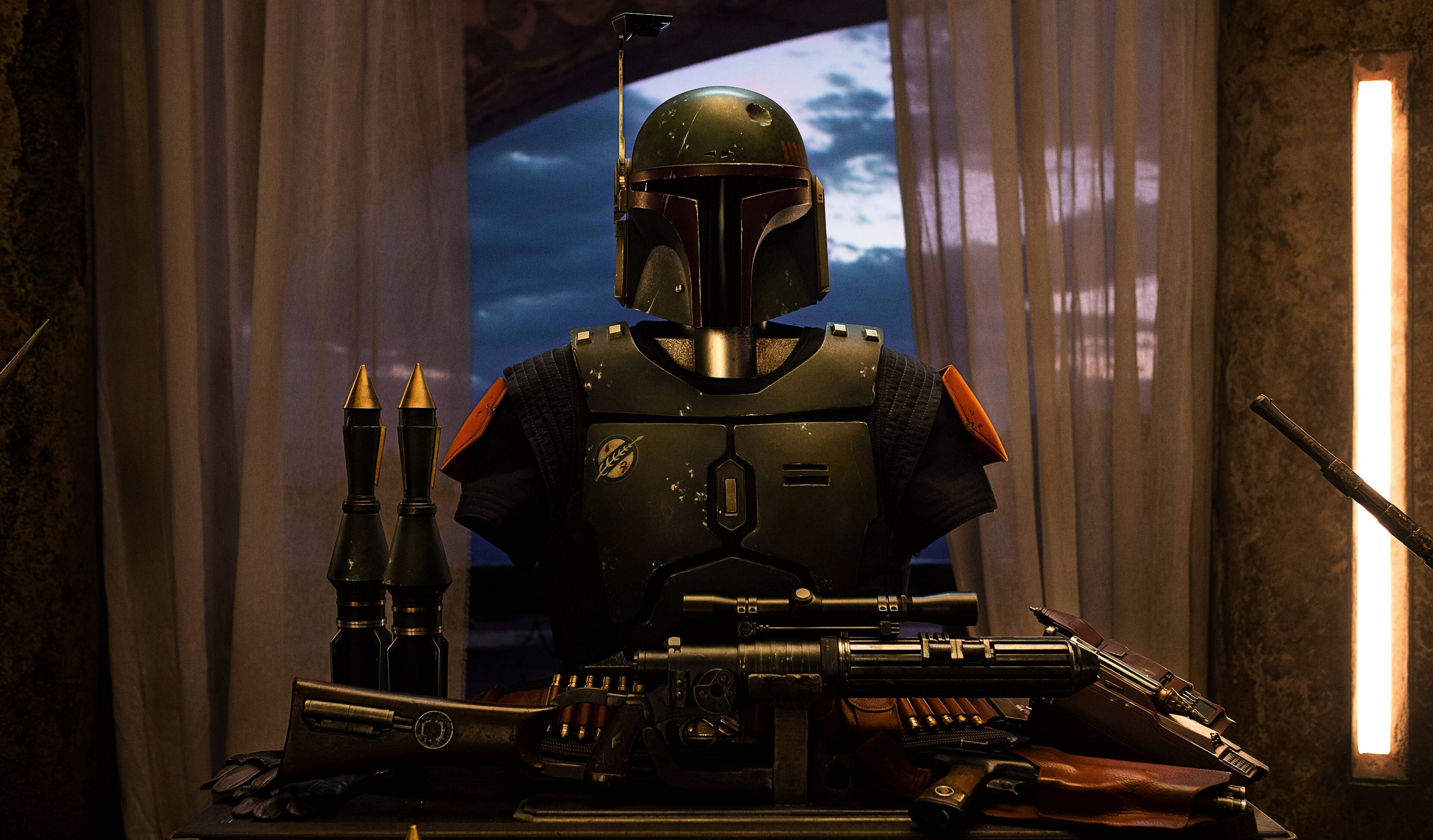 Boba 桌布 壁紙 — 一名身穿綠色盔甲的曼達洛戰士站在武器桌後面，背景是透過窗簾可見的暮色天空。