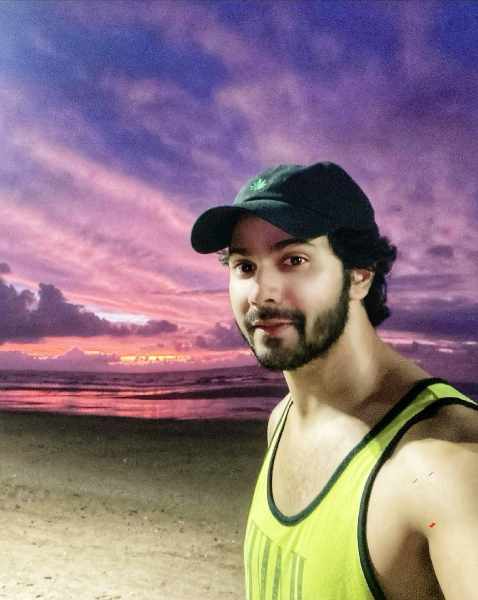 Varun 桌布 壁紙 — 一名身穿綠色背心和黑色鴨舌帽的男子站在日落時分的沙灘上，上方有鮮豔的紫色和粉紅色雲彩。