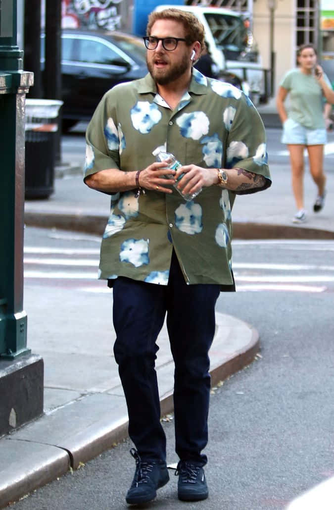 jonah hill 桌布 壁紙 — 一名身穿綠色花卉圖案襯衫的男子，戴著耳塞，拿著水瓶，在城市人行道上行走。