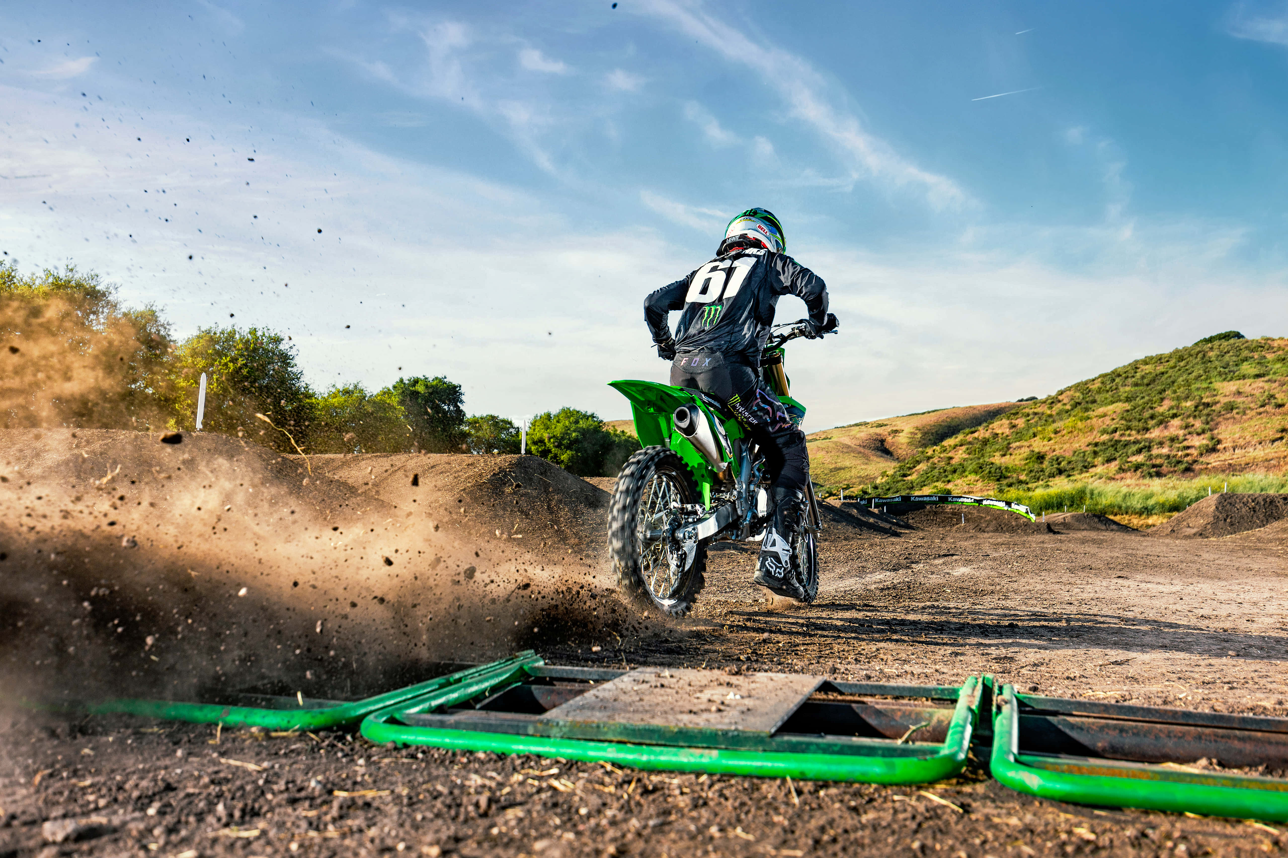 Dirtbike 桌布 壁紙 — 一名身穿綠色裝備的越野摩托車手在陽光明媚的賽道上飛越泥土跳台，塵土飛揚，背景是山丘。