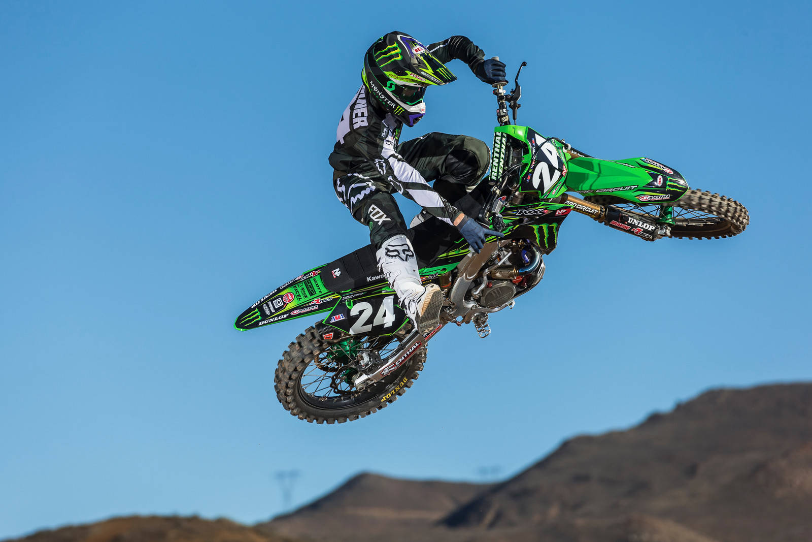 Motocross 桌布 壁紙 — 一名身穿綠色裝備的越野機車騎士在藍天和遠山背景下，騎著綠色機車在空中飛翔。