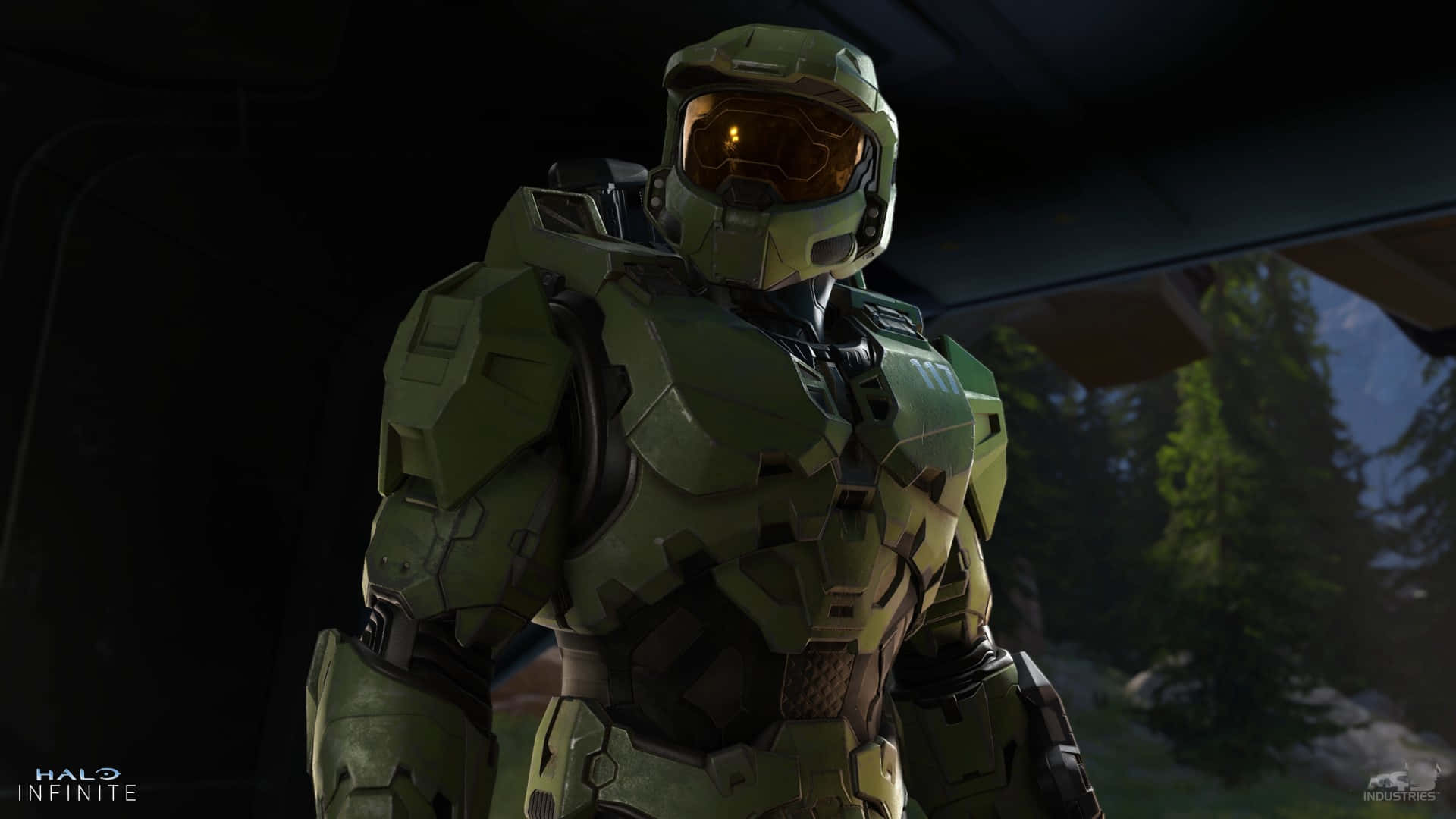 Halo Infinite 桌布 壁紙 — 一名身穿綠色裝甲服、戴著發光橙色護目鏡的士兵站在昏暗的室內，外面可見森林。