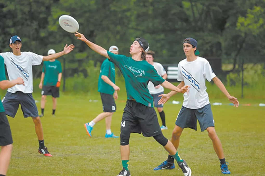 Frisbee 桌布 壁紙 — 一名身穿綠色襯衫的年輕男子跳起接住白色飛盤，隊友和對手在草地上專注觀看。