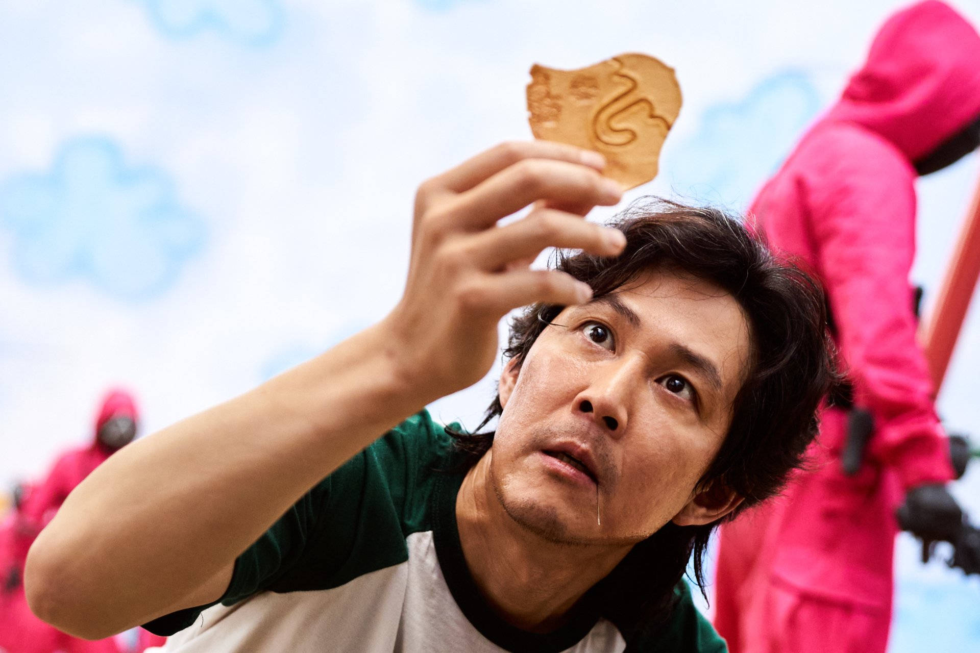 Actors 桌布 壁紙 — 一名身穿綠色襯衫的男子，在陰天的天空下舉起一個帶有蛇形圖案的木製代幣，背景有粉紅色西裝的人物。