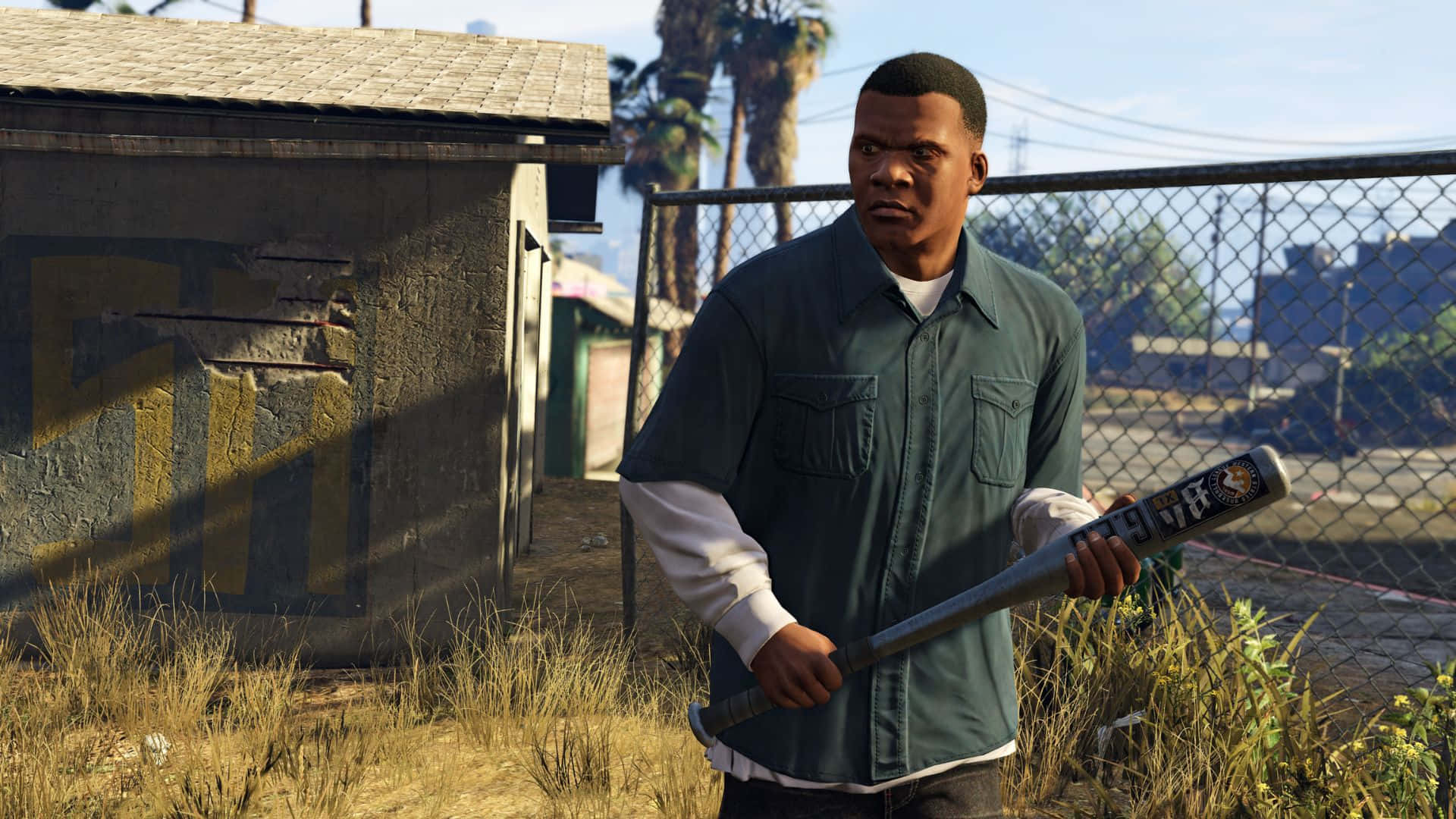Grand Theft Auto V 桌布 壁紙 — 一名身穿綠色襯衫的男子在陽光明媚的城市庭院中，靠近鐵絲網圍欄，手持棒球棒，周圍有棕櫚樹。