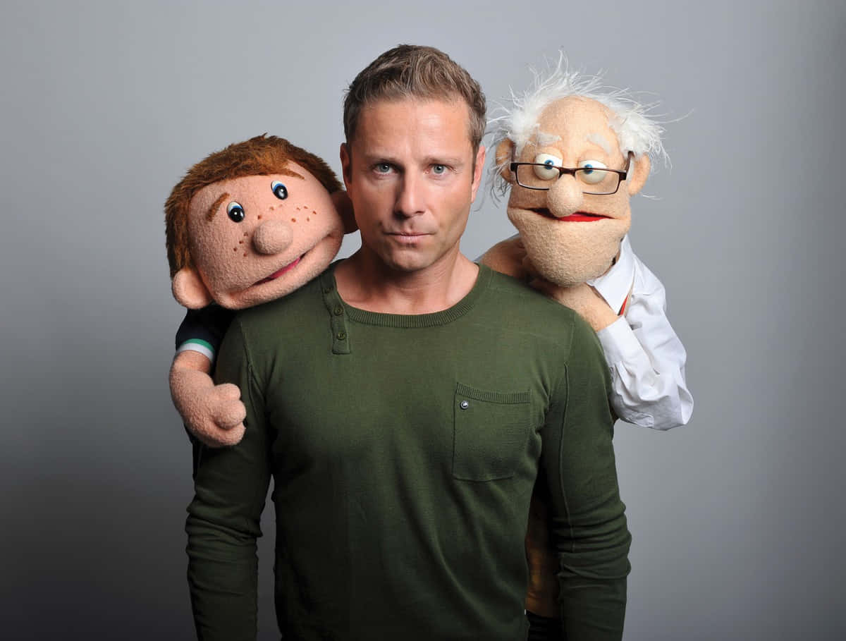 puppet 桌布 壁紙 — 一名身穿綠色襯衫的男子站在中央，肩上放著兩個木偶——一個有雀斑，一個戴眼鏡——背景是純灰色。