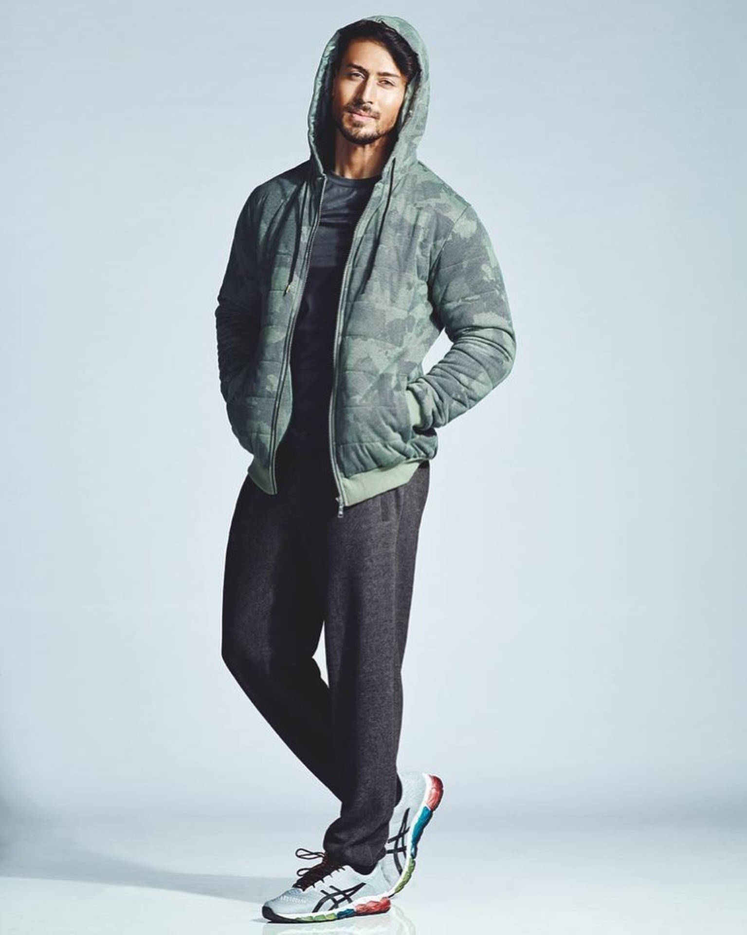 Tiger Shroff 桌布 壁紙 — 一名身穿綠色迷彩連帽外套和深色運動褲的男子，自信地站在純色淺灰色攝影棚背景前。