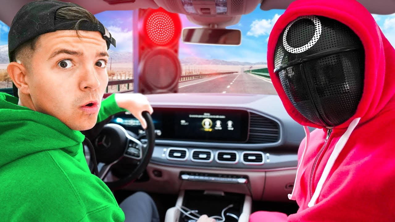 Squid Game 桌布 壁紙 — 一名身穿綠色連帽衫的男子震驚地凝視著，一名戴著面具、身穿紅色連帽衫的人坐在他旁邊，在一輛停在紅燈前的豪華車內。