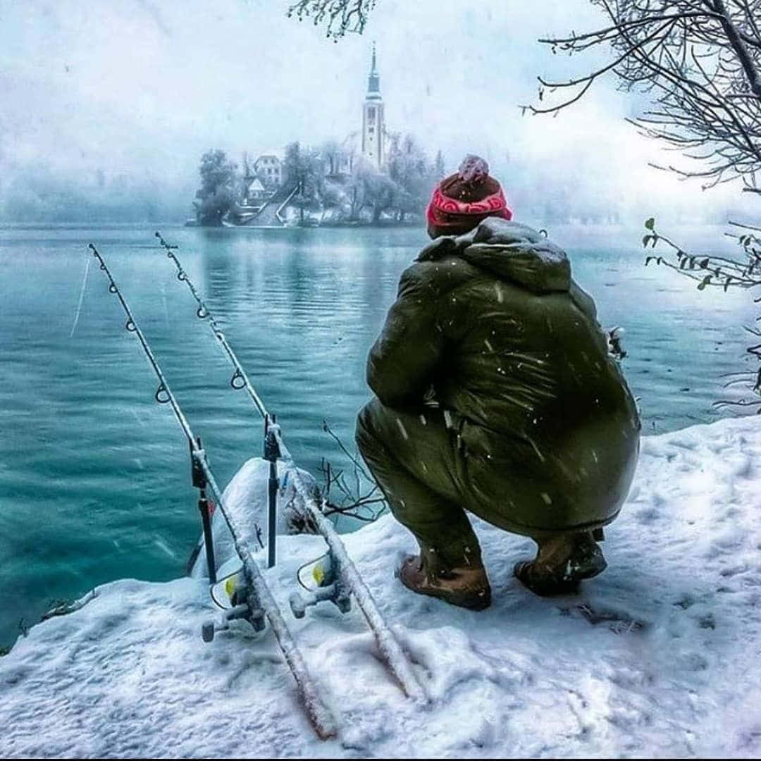 Ice Fishing 桌布 壁紙 — 一名身穿綠色雪衣和紅色毛帽的人蹲在積雪的湖岸邊釣魚，雪花飄落在遠處的島嶼教堂周圍。