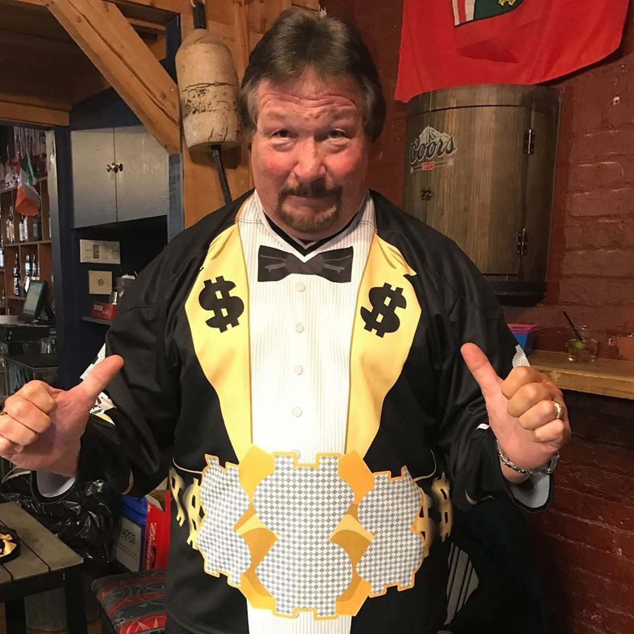 WWE 桌布 壁紙 — 一名身穿美元符號主題燕尾服的男士，在一個有磚牆和Coors啤酒桶的鄉村風格酒吧裡豎起兩個大拇指。