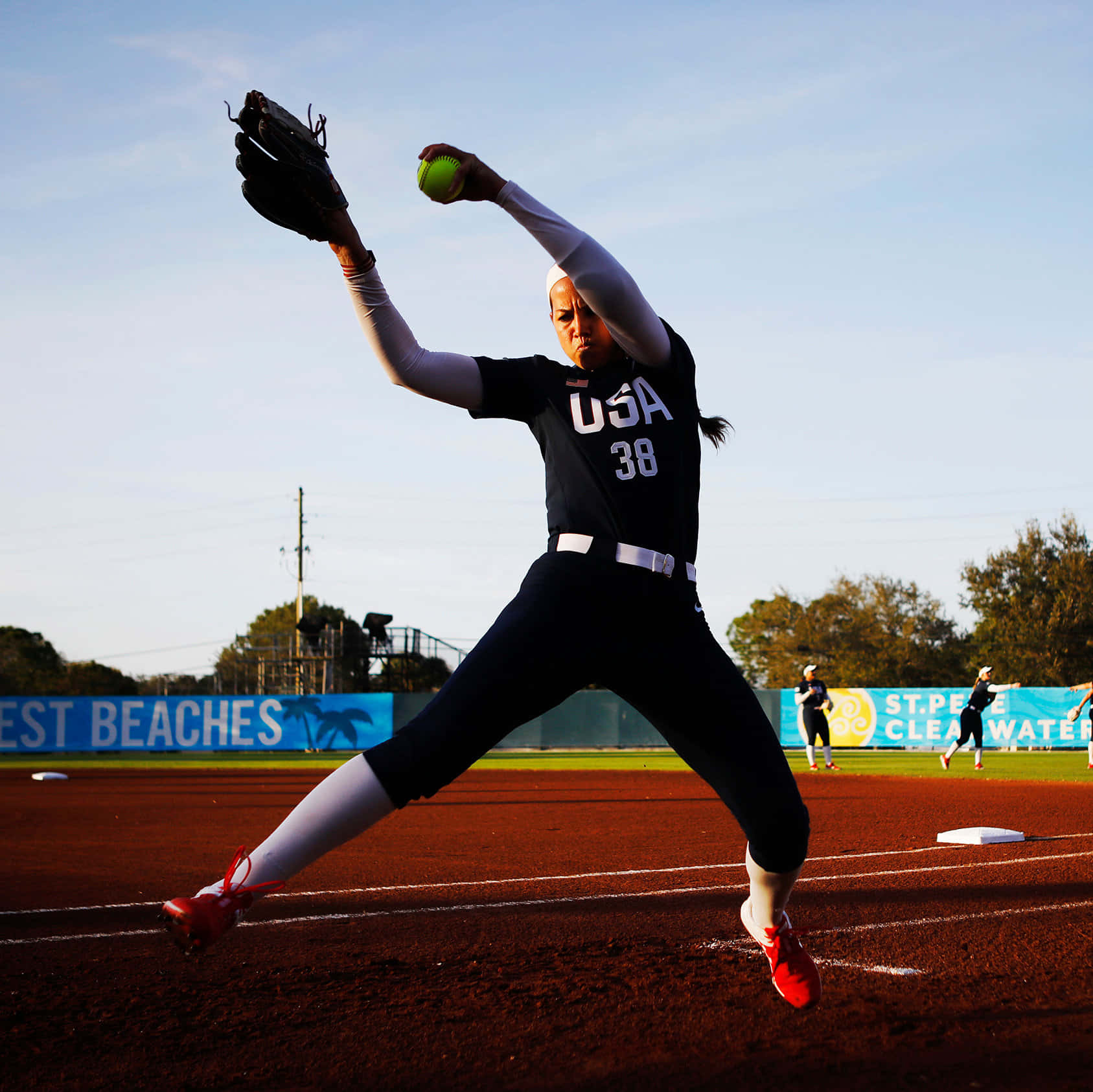 Softball 桌布 壁紙 — 一名身穿美國球衣的女壘球投手在陽光明媚的球場上投出強力一球，背景有隊友。
