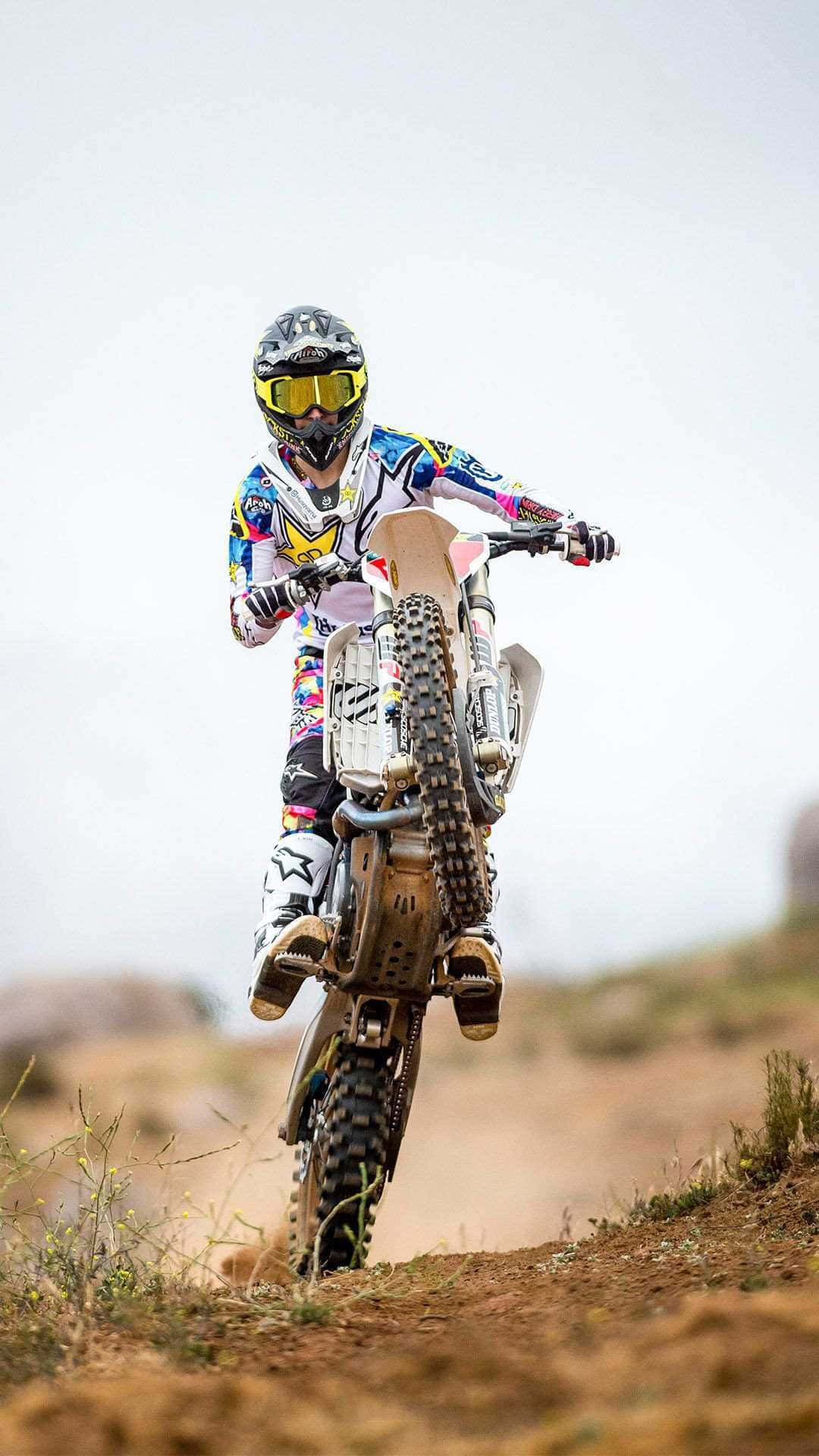 Dirtbike 桌布 壁紙 — 一名身穿色彩繽紛裝備的越野摩托車手，在多雲天空下的塵土小徑上傾斜入彎。
