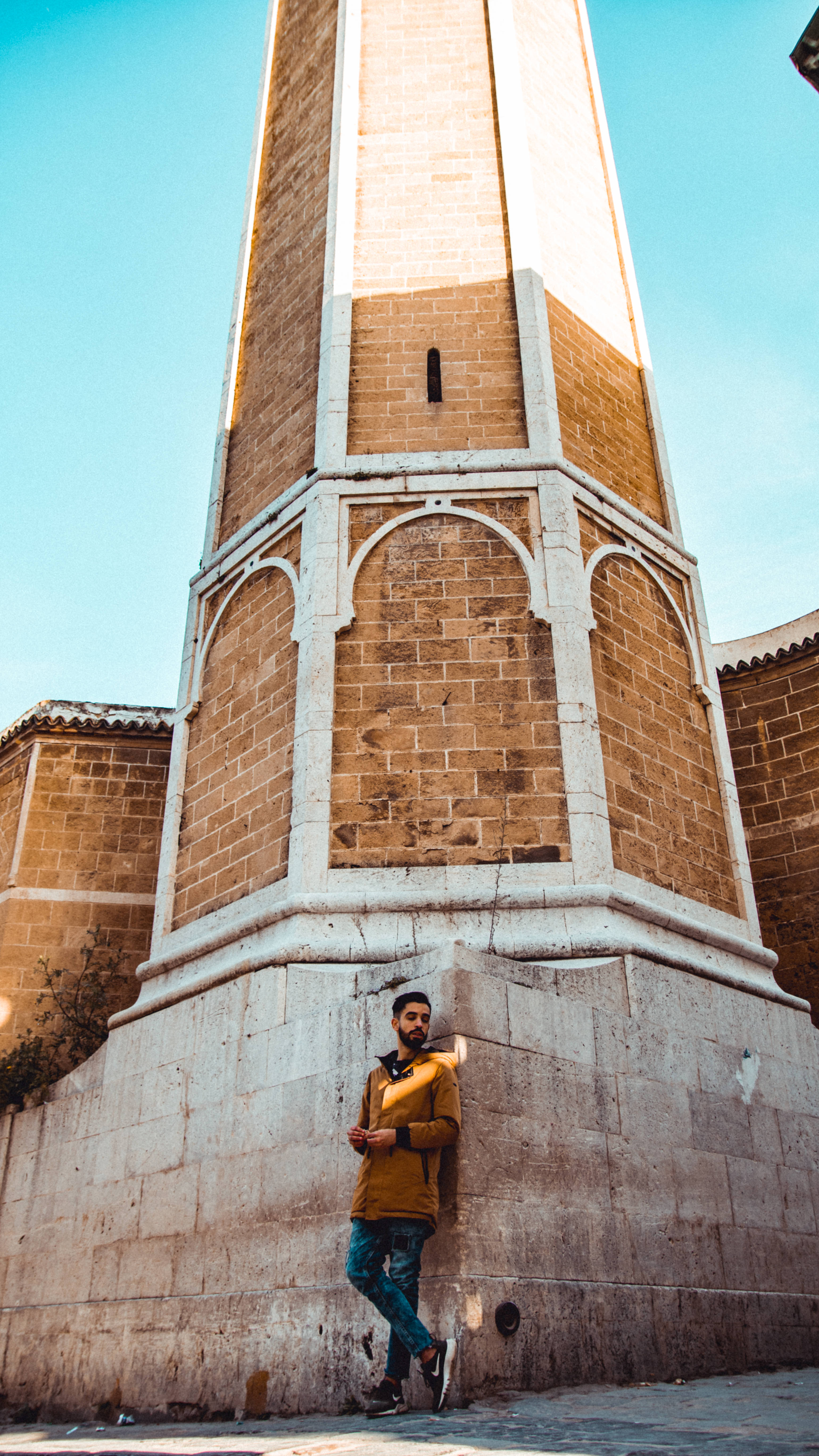 Tunisia 桌布 壁紙 — 一名身穿芥末黃色外套的年輕男子，在晴朗的藍天下，隨意地靠在高聳宣禮塔的古老石基上。