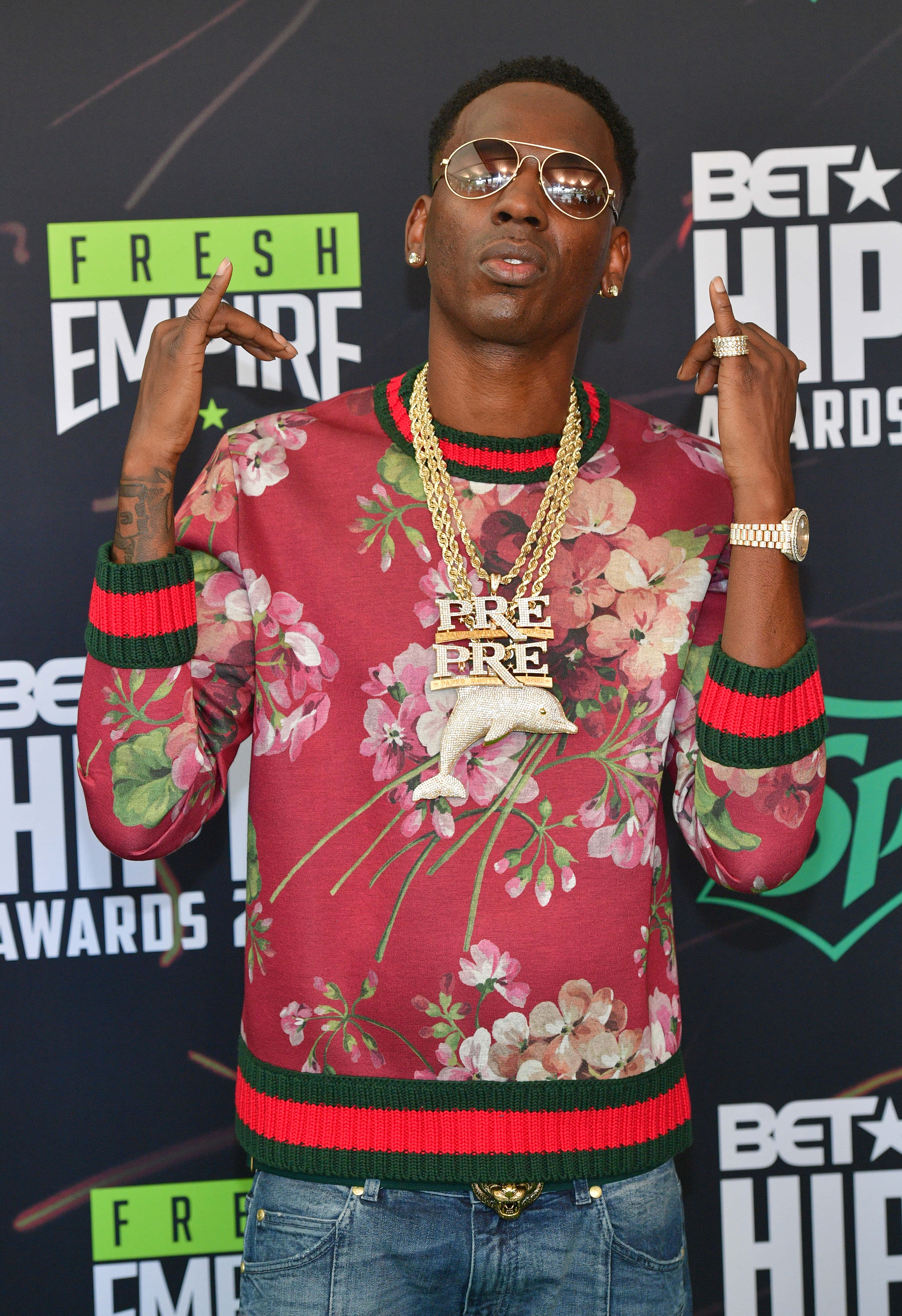 Young Dolph 桌布 壁紙 — 一名身穿花卉圖案紅色毛衣和金色鏈條的男子，在BET嘻哈頒獎典禮上戴著太陽眼鏡並做出手指手勢，自信地擺姿勢。