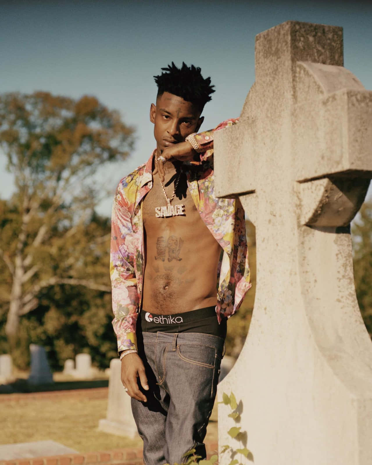 21 Savage 桌布 壁紙 — 一名身穿花卉圖案襯衫的男子，在陽光明媚的墓園裡靠著一塊風化的墓碑，背景有樹木。