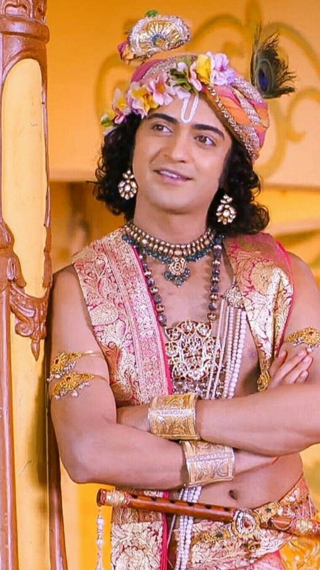 radha krishna serial 桌布 壁紙 — 一名身穿華麗傳統印度服飾、頭戴花冠、佩戴黃金首飾的男子，在金色背景前自信地交叉雙臂站立。