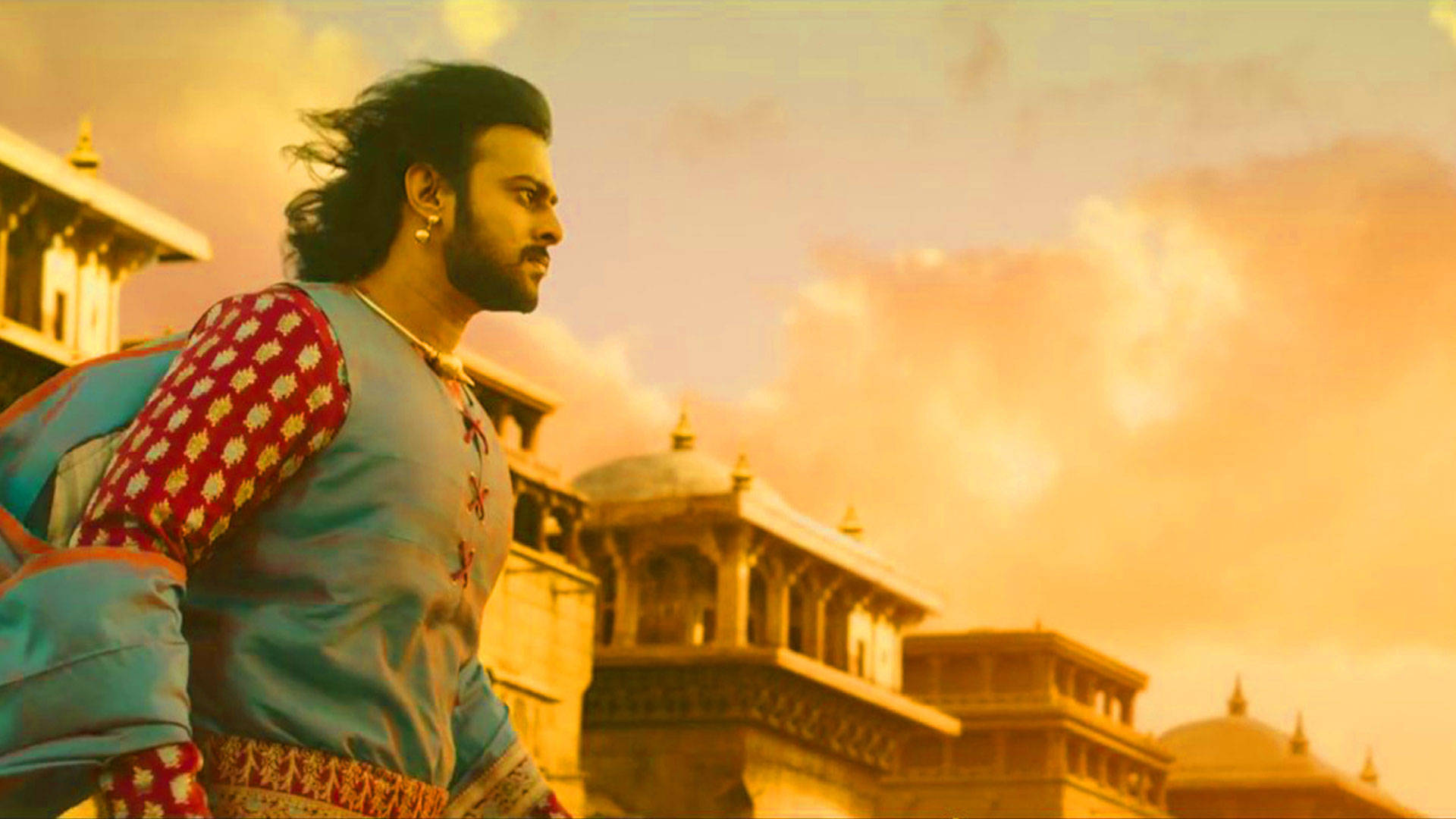 Prabhas HD 桌布 壁紙 — 一名身穿華麗傳統服飾的男子，在溫暖的金色夕陽天空下，站在古老的圓頂建築前。
