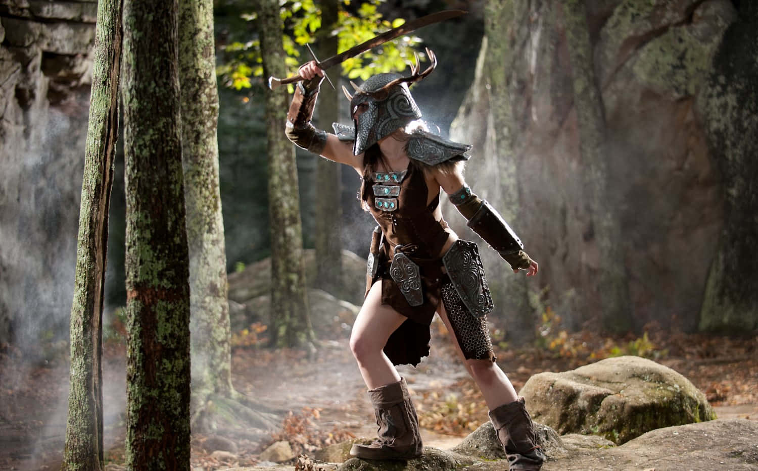 Aela The Huntress 桌布 壁紙 — 一名身穿華麗盔甲和角盔的戰士，在迷霧森林中，周圍是長滿苔蘚的樹木和落葉，手持劍擺出動態姿勢。