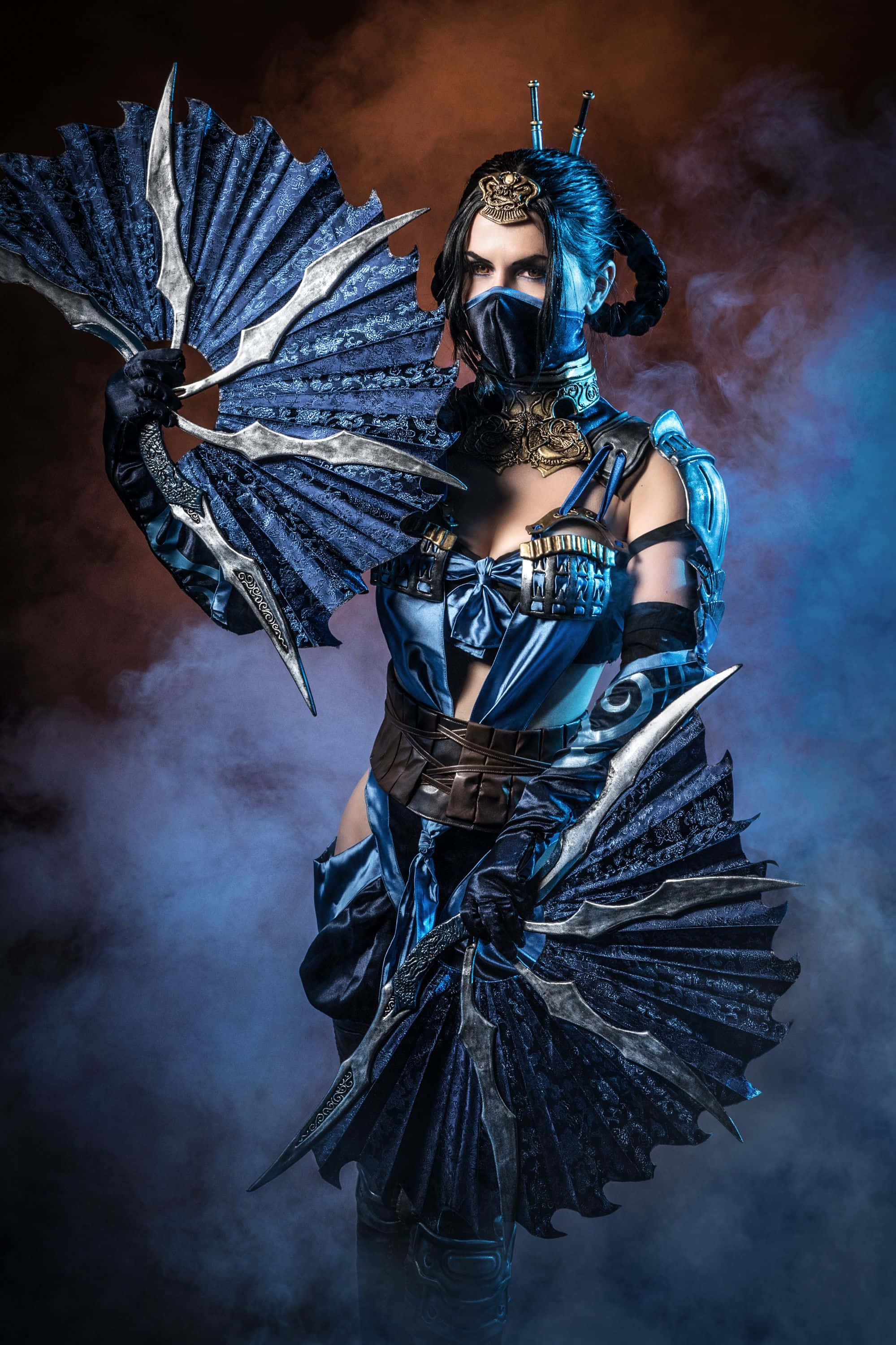 Mortal Kombat Cosplays 桌布 壁紙 — 一名身穿華麗藍色盔甲的戰士，在煙霧繚繞的暮色背景下揮舞雙扇刃。