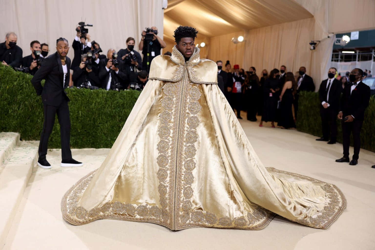 Met Gala 2021 桌布 壁紙 — 一名身穿華麗金色刺繡斗篷長袍的男子自信地走在紅地毯上，兩旁是攝影師和綠色樹籬。