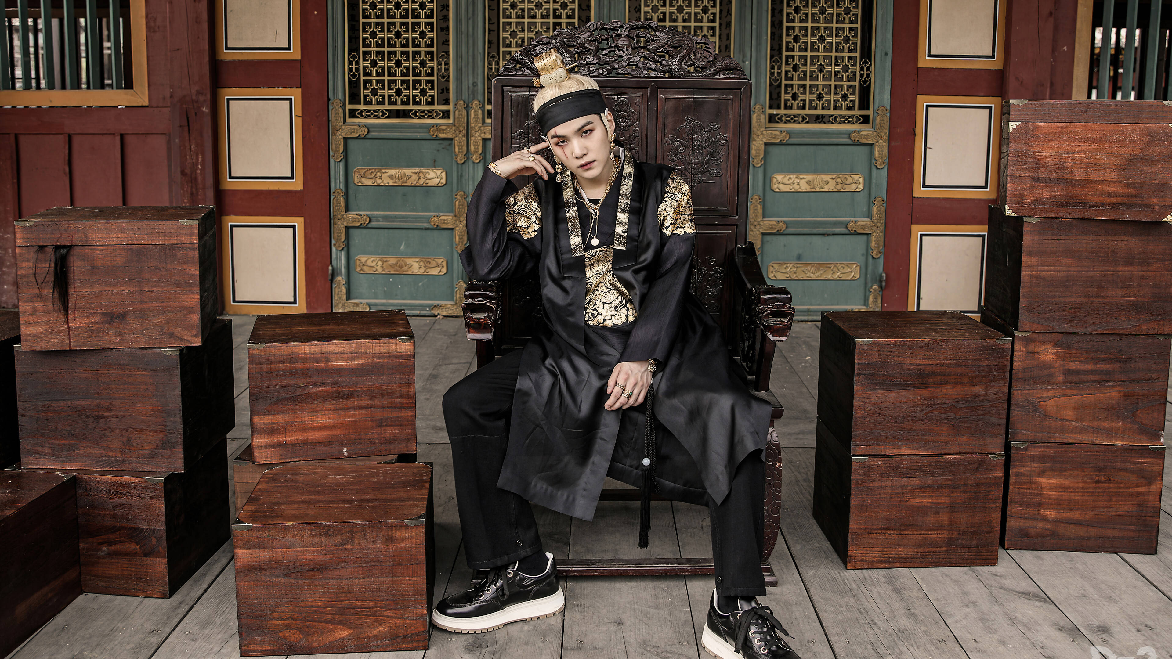 Suga 桌布 壁紙 — 一名身穿華麗黑金傳統服飾的男子，威嚴地坐在歷史建築內堆疊木箱環繞的古董木製王座上。